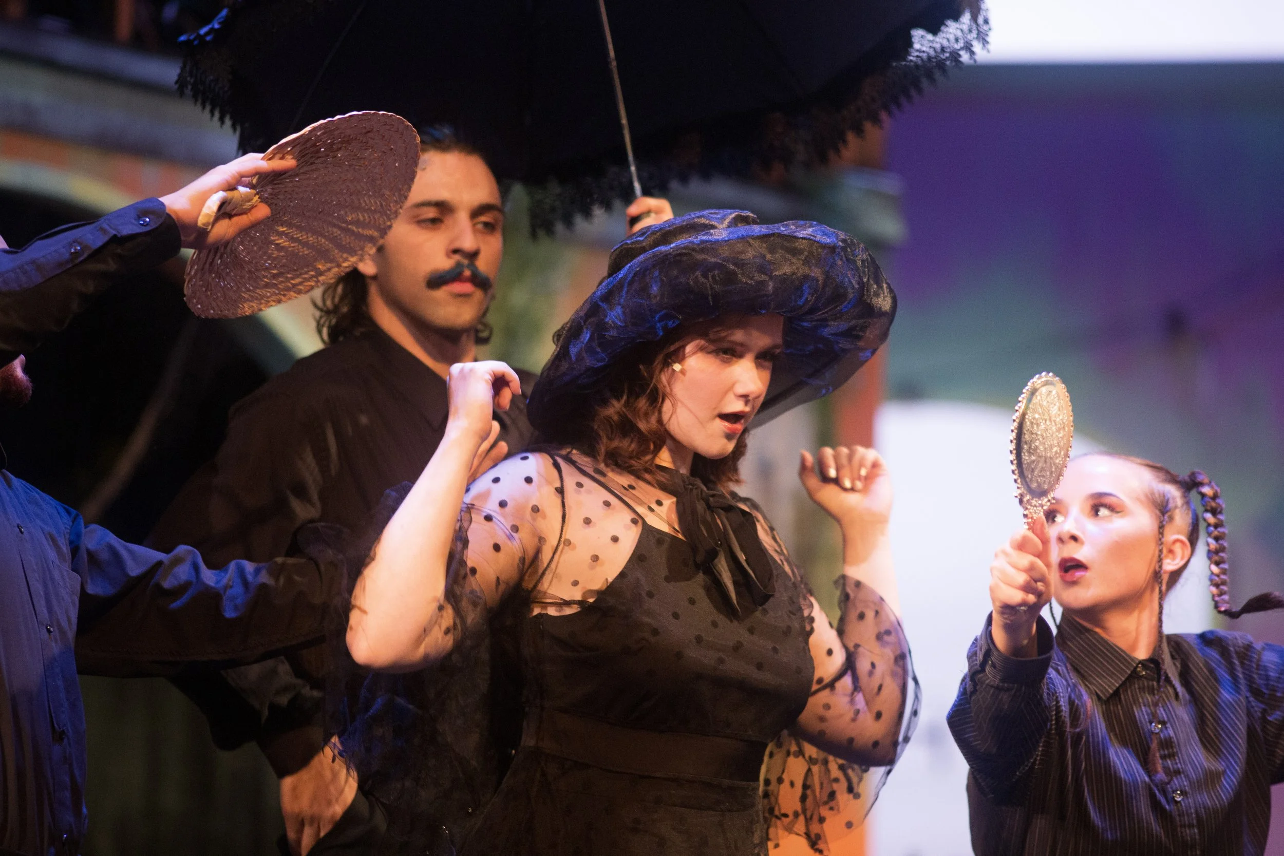 Three women and two men in theatrical costumes on stage, with one woman adjusting her hat and another looking into a mirror.
