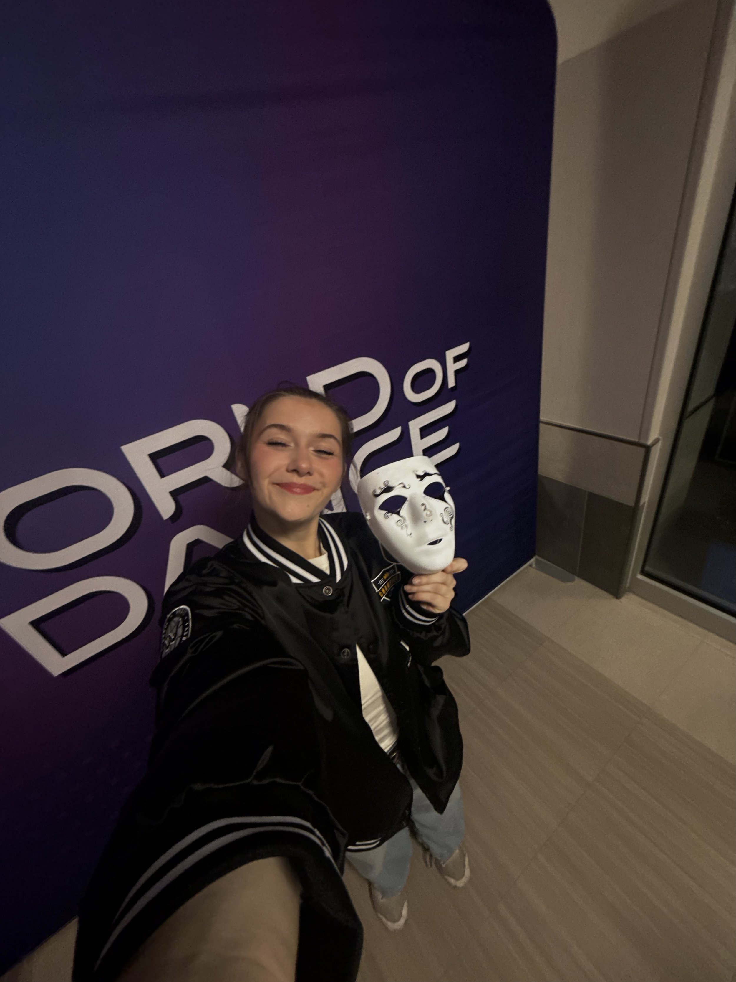 A young woman taking a selfie while holding a white mask, standing in front of a purple wall with the words 'World of' Dance visible.