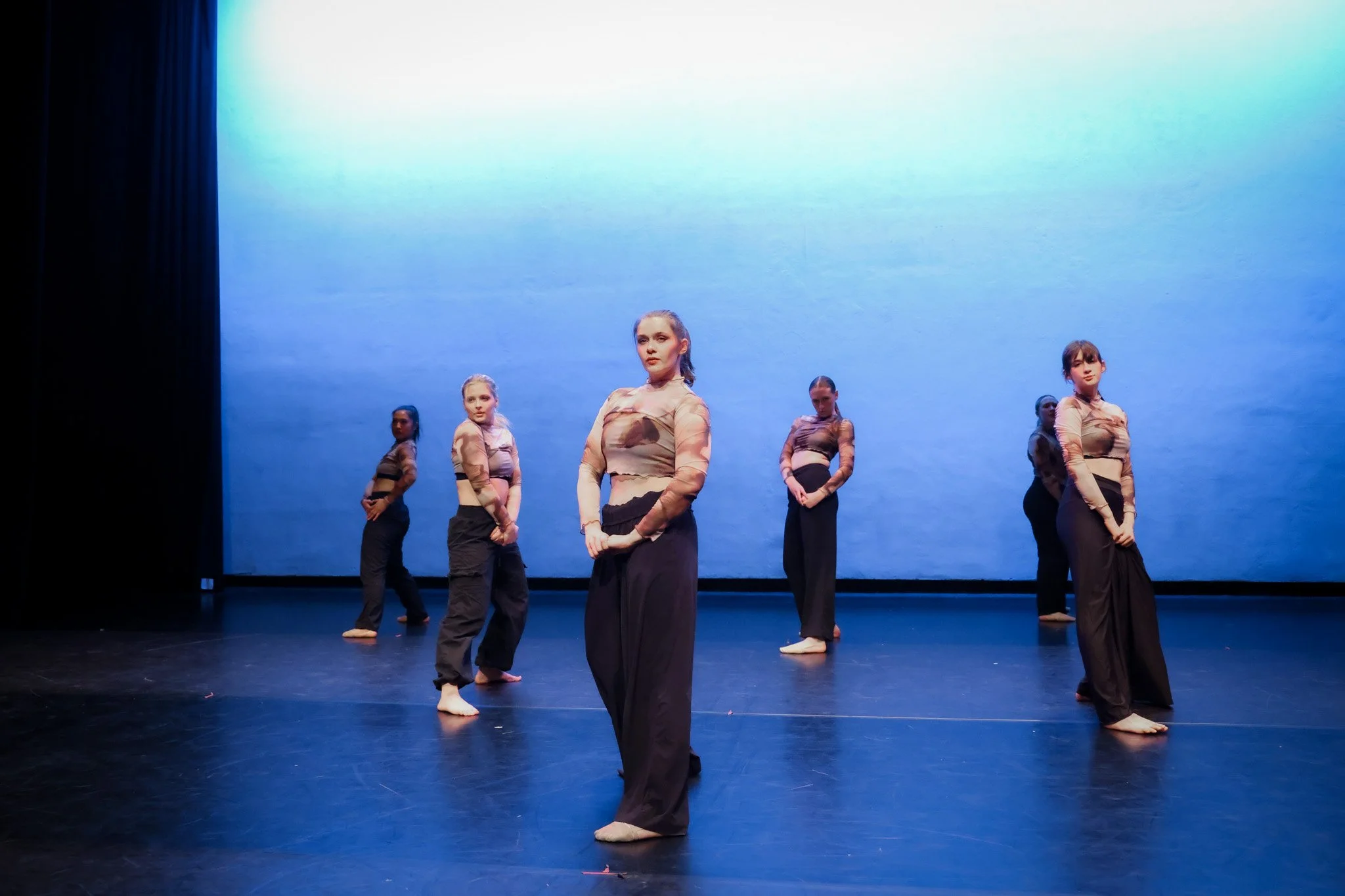 Five young women in dance costumes stand on stage with a blue background, practicing a dance routine.