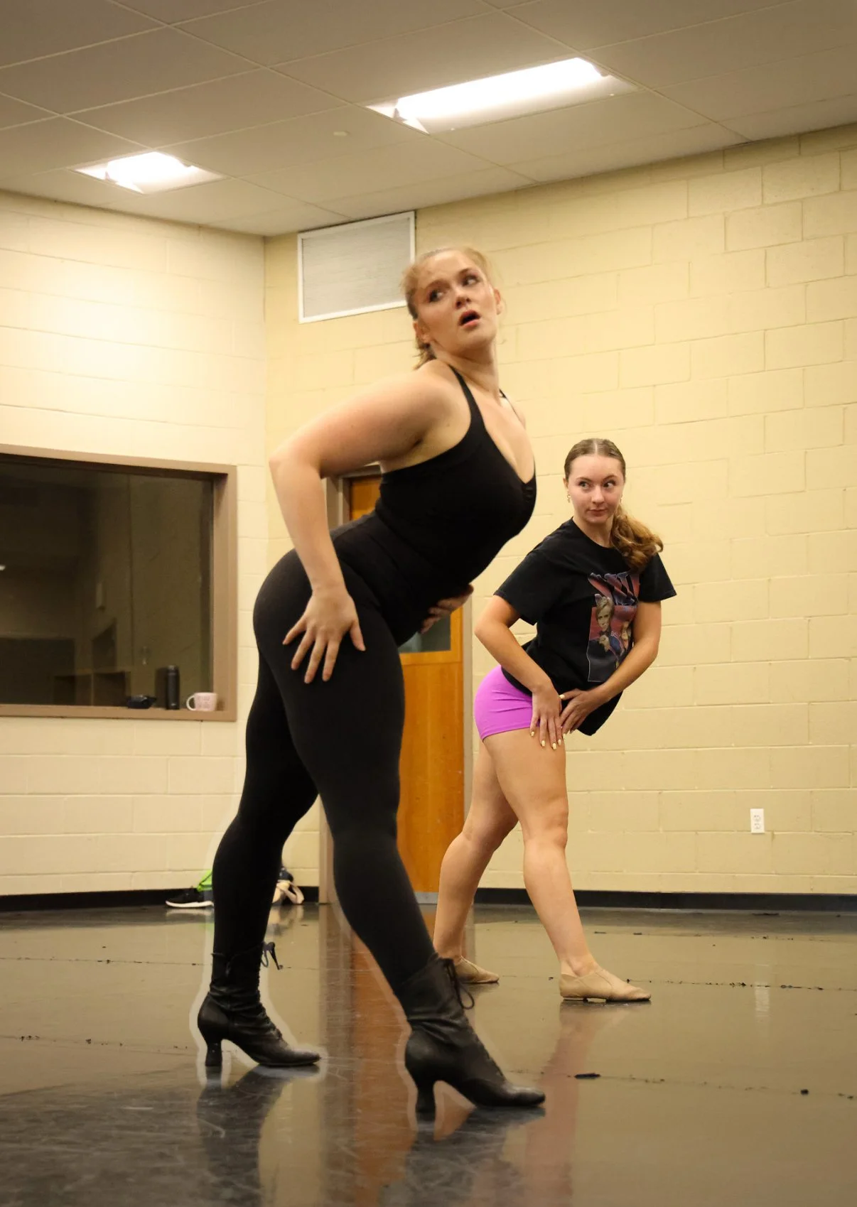 Two women practicing dance in a studio, one in black leggings and boots, the other in pink shorts and a black T-shirt, both in dance poses.
