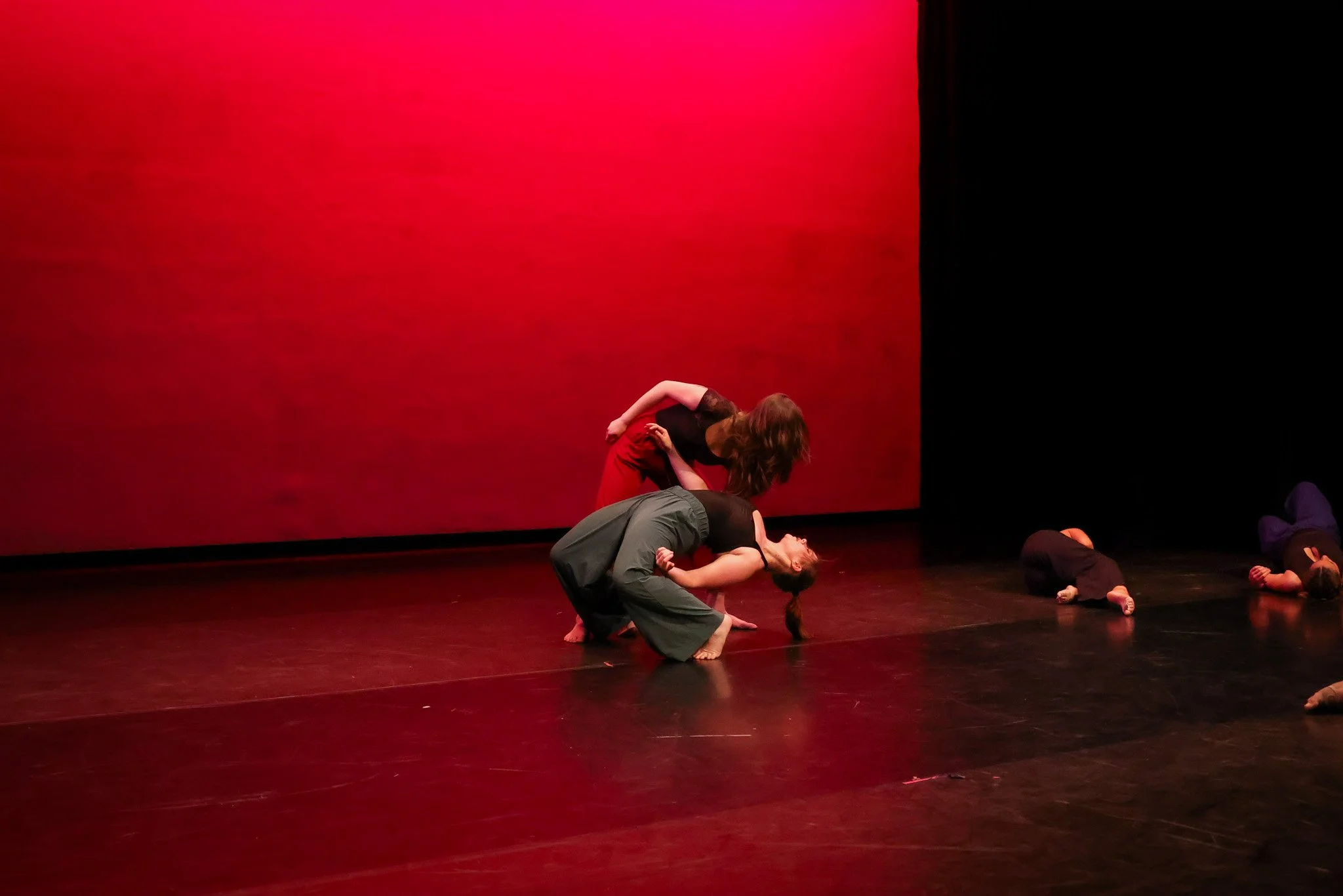 Dance performance on stage with a red background, featuring two women in a dramatic pose.