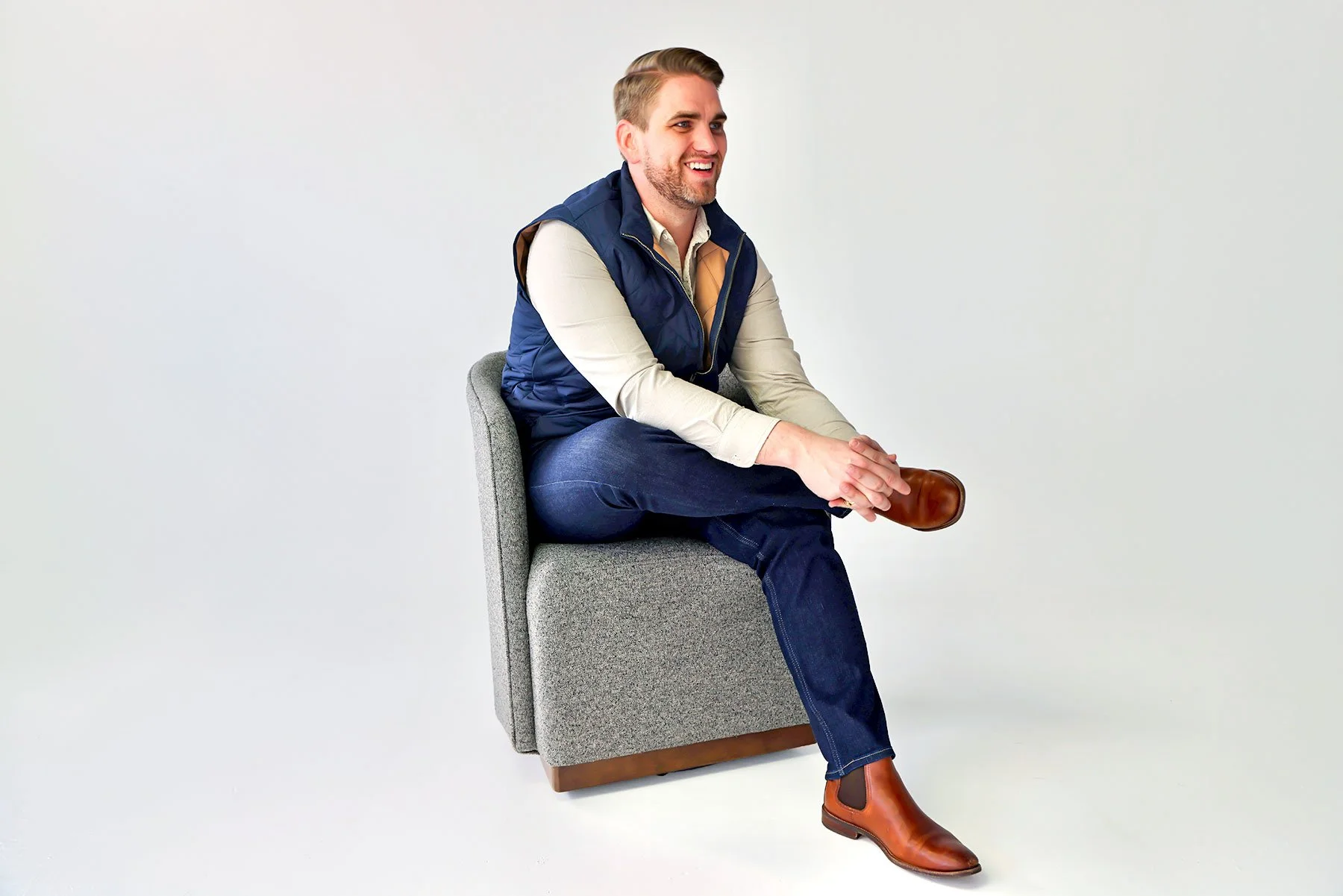 A man sitting on a gray chair, wearing a beige shirt, navy vest, jeans, and brown boots, smiling and holding his ankle.