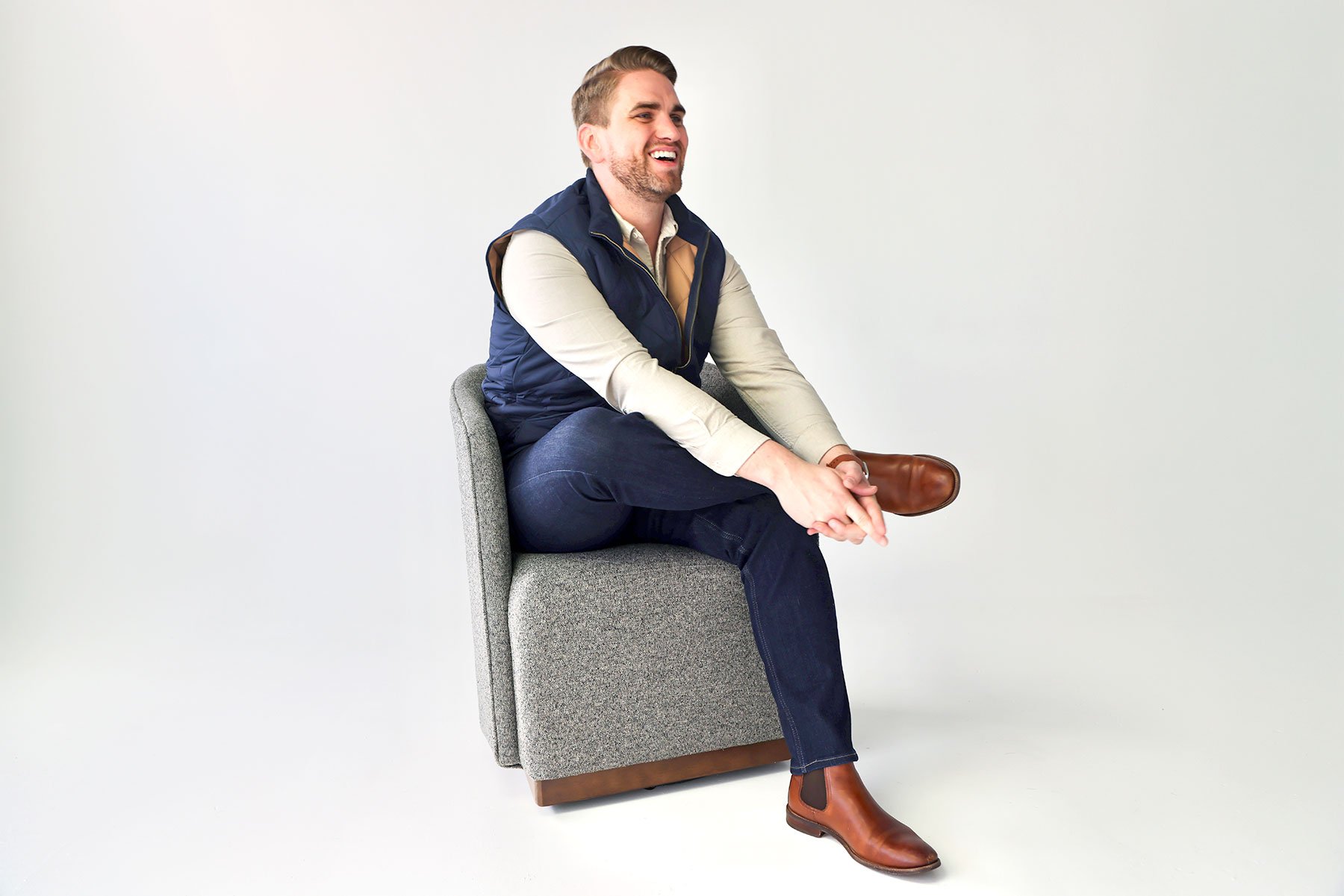 A man sitting on a gray armchair, laughing, wearing a beige shirt, dark blue vest, dark jeans, and brown boots, against a plain white background.