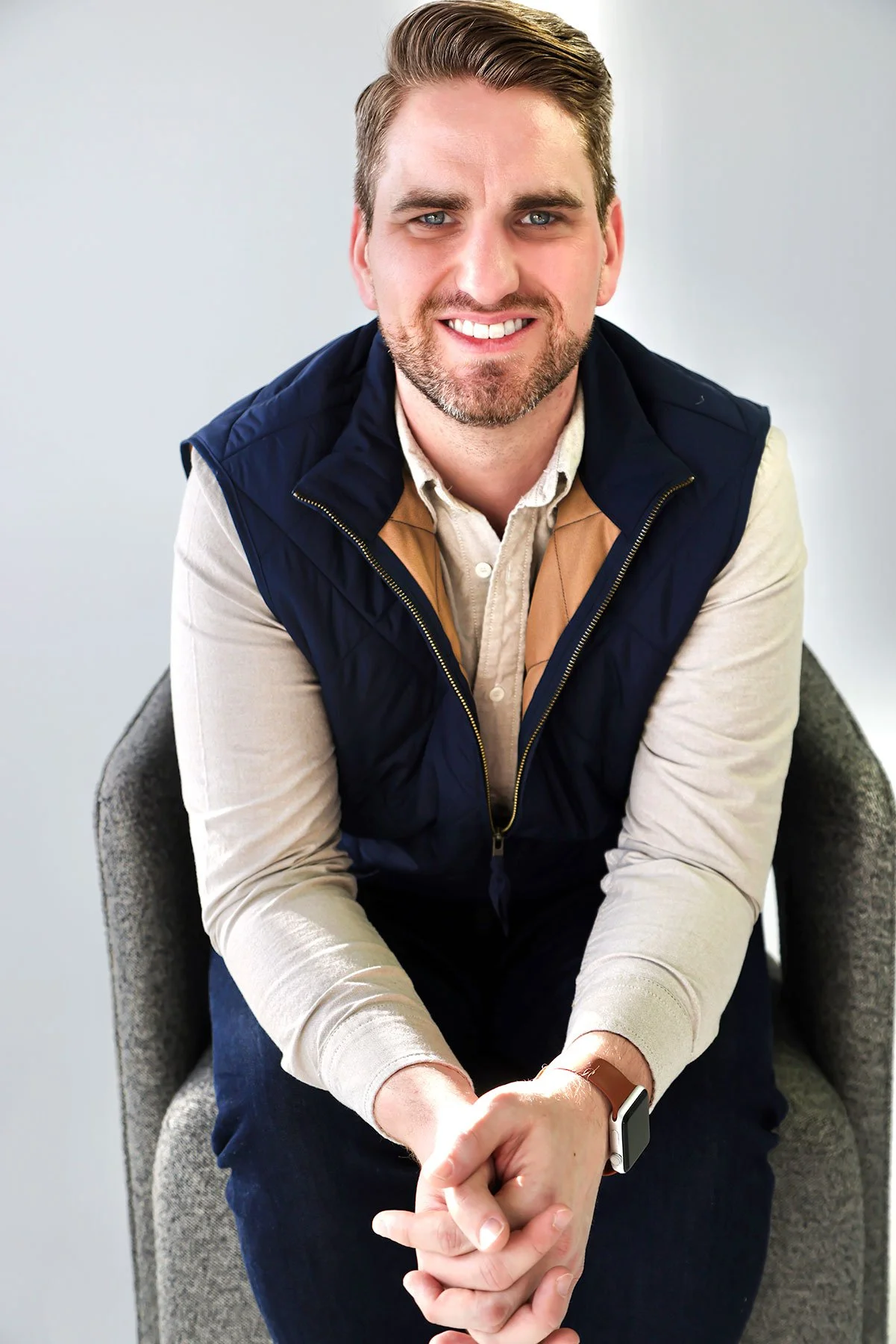 A smiling man with brown hair and a beard, wearing a beige shirt and a dark blue vest, sitting on a gray chair with his hands clasped.