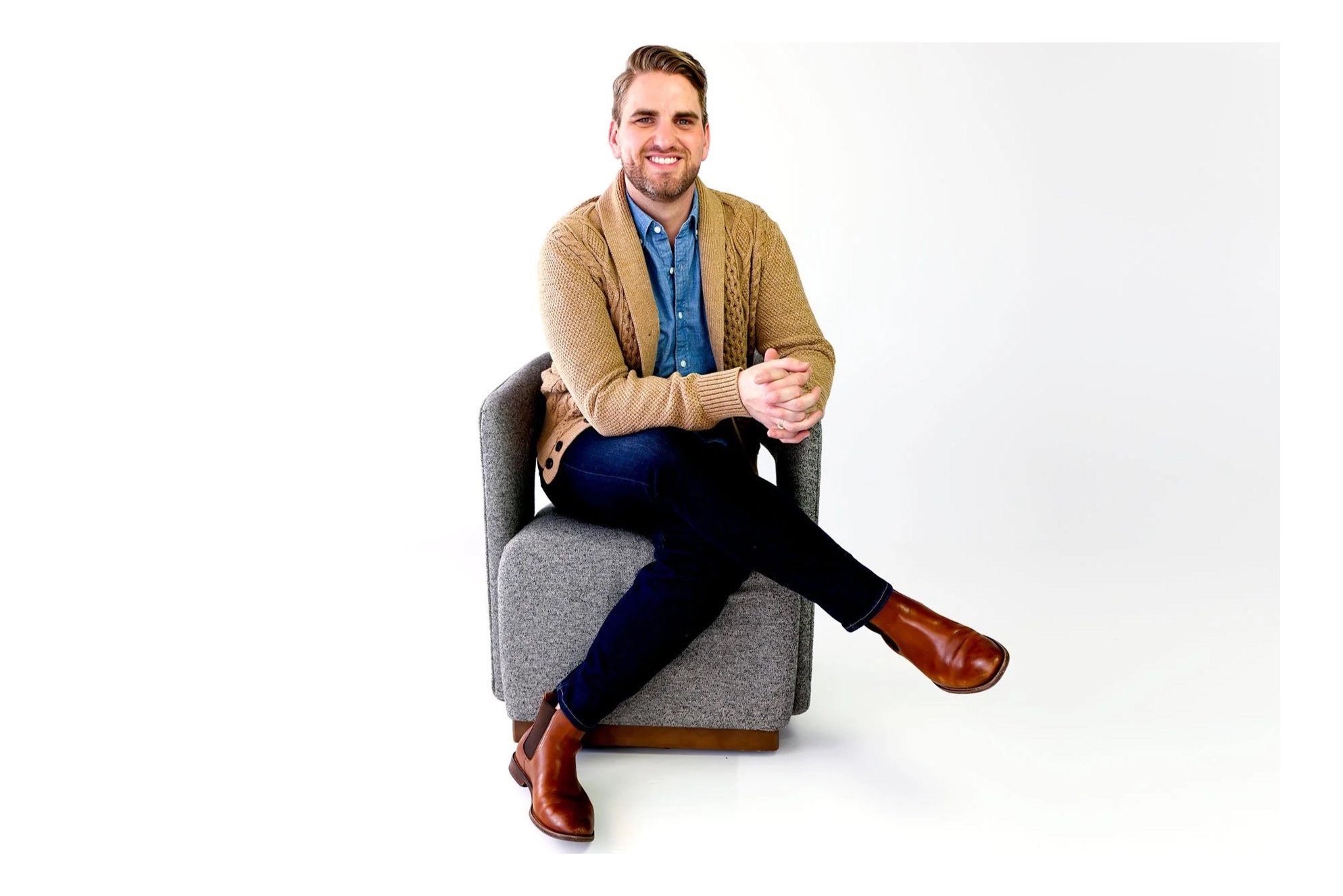 A man sitting on a gray armchair, smiling, wearing a tan cardigan, blue shirt, dark pants, and brown boots, against a white background.