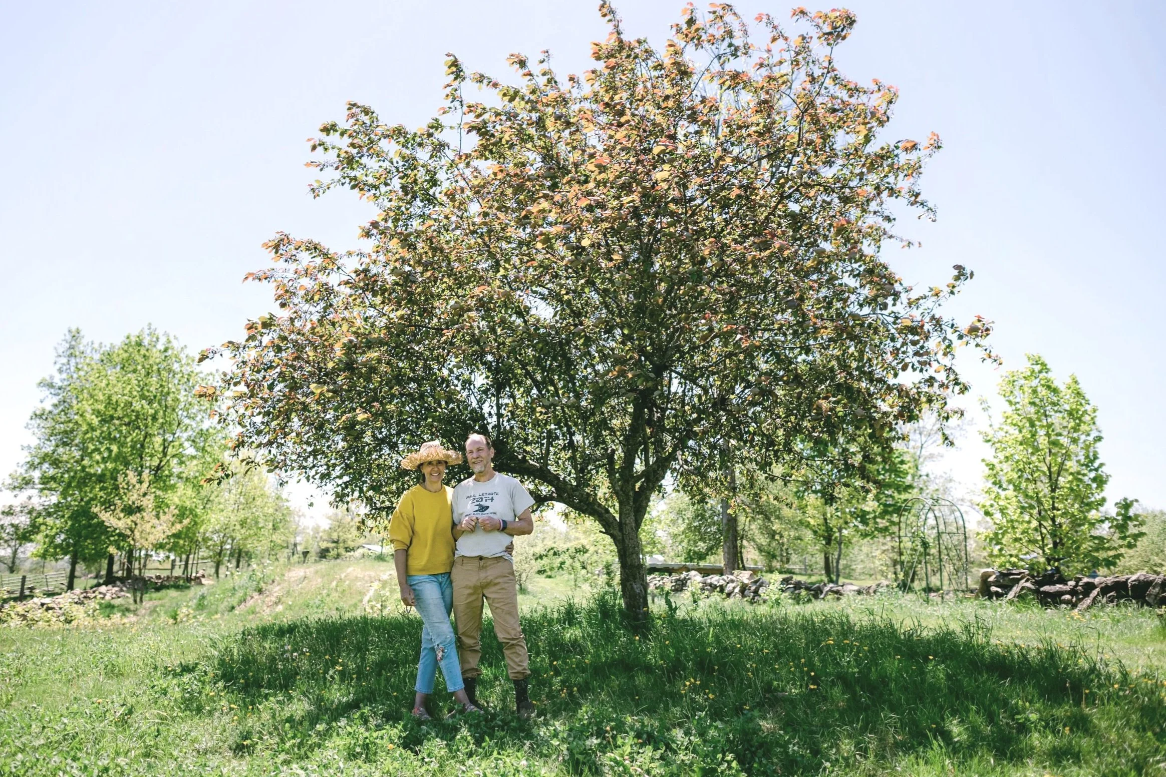 A smiling woman (Daniela -owner) in a yellow sweater and a sun hat, and a man (Patrick - owner) in a white T-shirt and work pants, standing under a large blooming crabapple tree on a sunny day on the farm