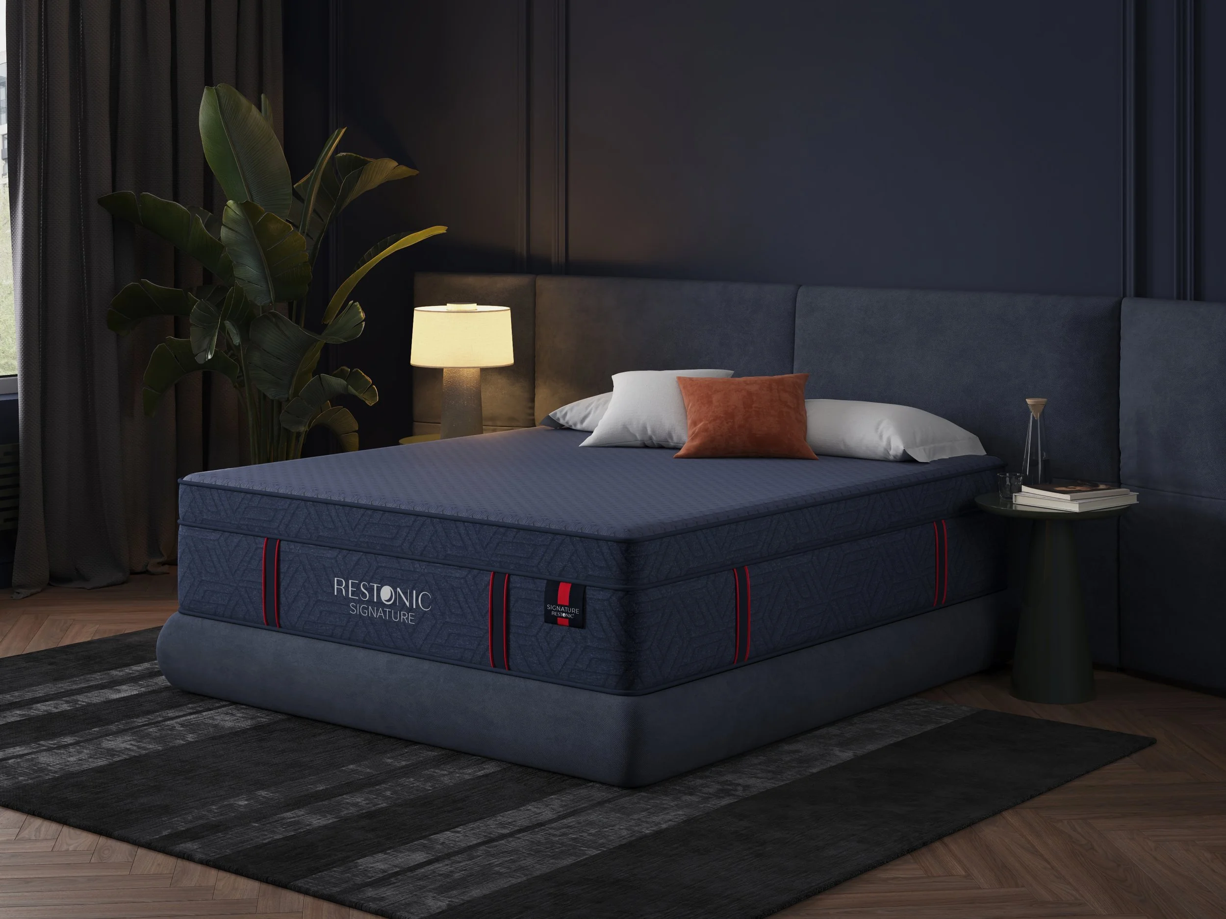 A modern bedroom with a Comfortably Restonic mattress on a bed frame, flanked by side tables with lamps, a large green plant, dark curtains, and a black and gray rug, with a dark paneled wall in the background.