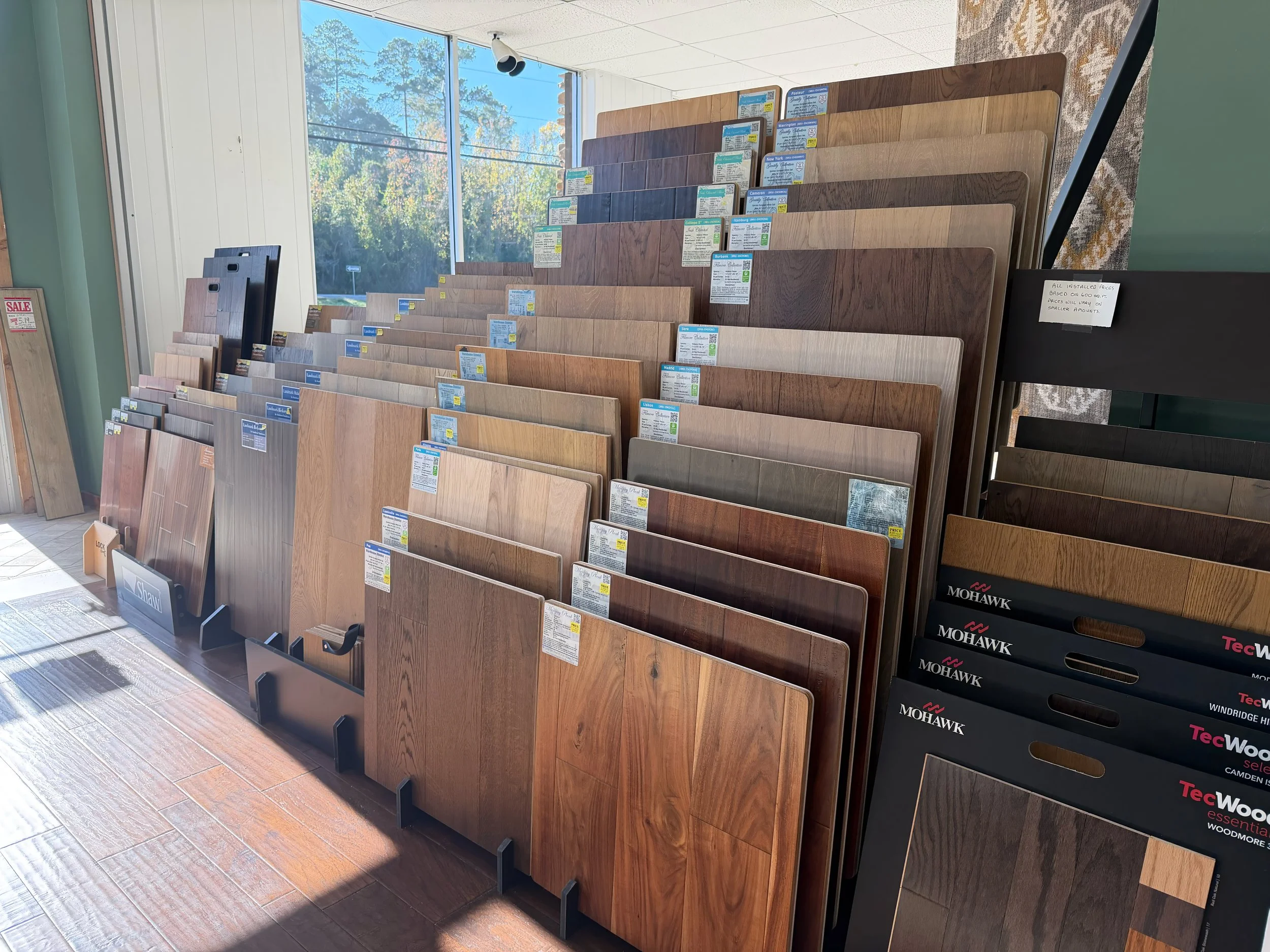 Display of wood flooring samples in a showroom, arranged on angled racks with labels, near a window with trees visible outside.