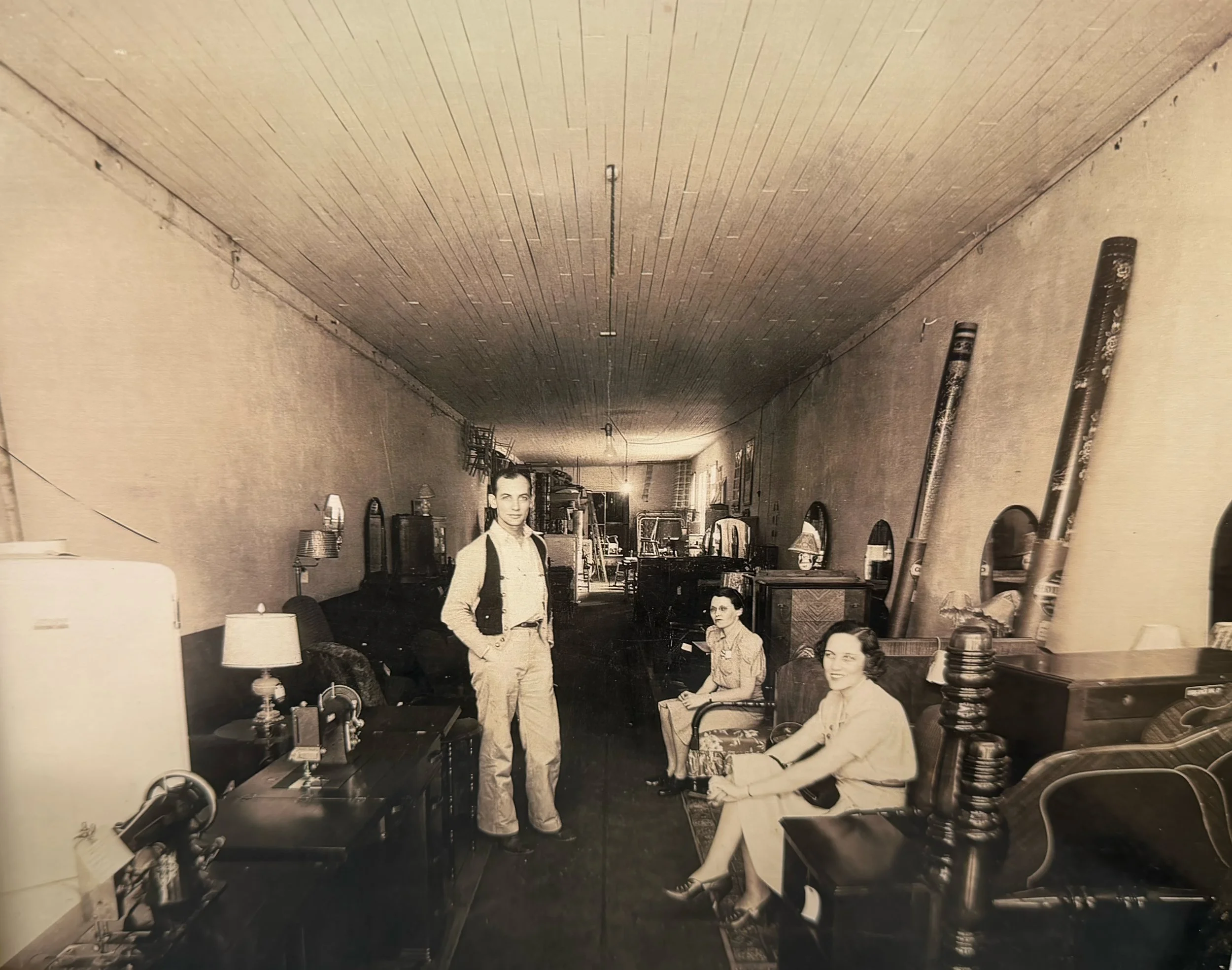 A vintage black-and-white photo of three people inside a sparsely furnished room with mirrors on the walls, two women sitting on chairs, a man standing next to a table, and baseball bats leaning against the wall.