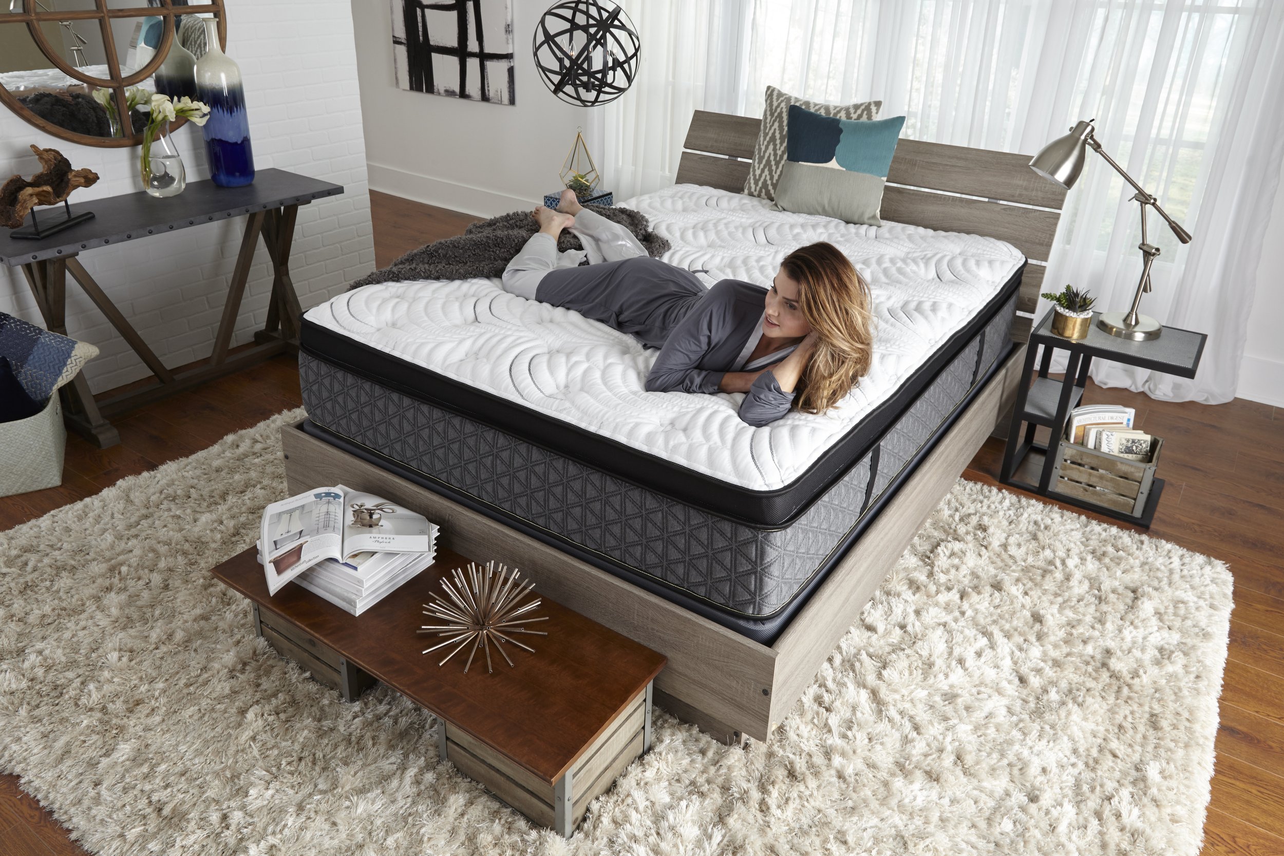 A woman lying on her side on a bed in a modern bedroom with white curtains, a textured white rug, and contemporary furniture.
