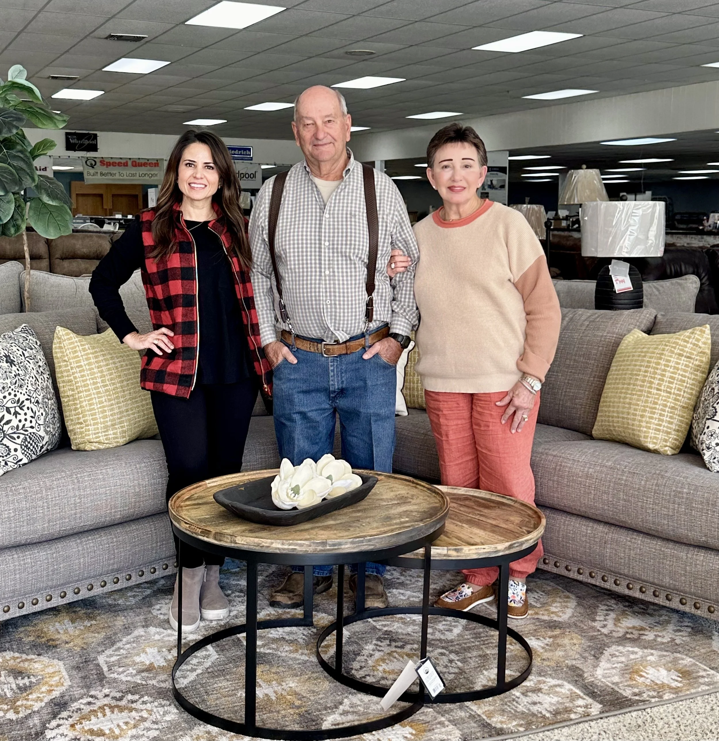 Three people, two women and one man, standing in a furniture store in front of a sofa with cushions. There are two round wooden tables with a decorative white sculpture on top. The store is well-lit and has various furniture items in the background.