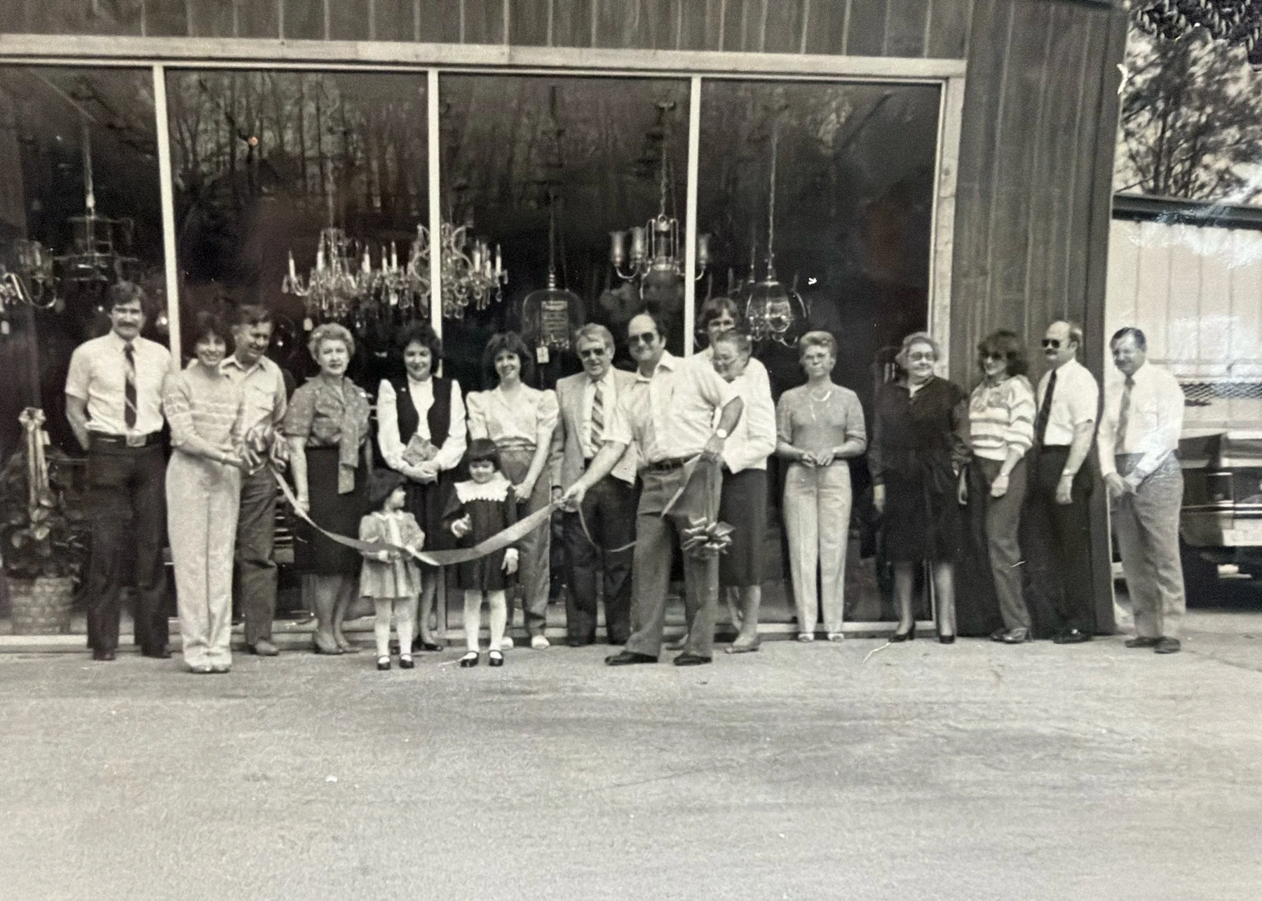 A group of people gathered outside a store with glass windows, possibly for a ribbon-cutting ceremony, with some holding a ribbon and scissors, in a black-and-white photograph likely from the mid-20th century.