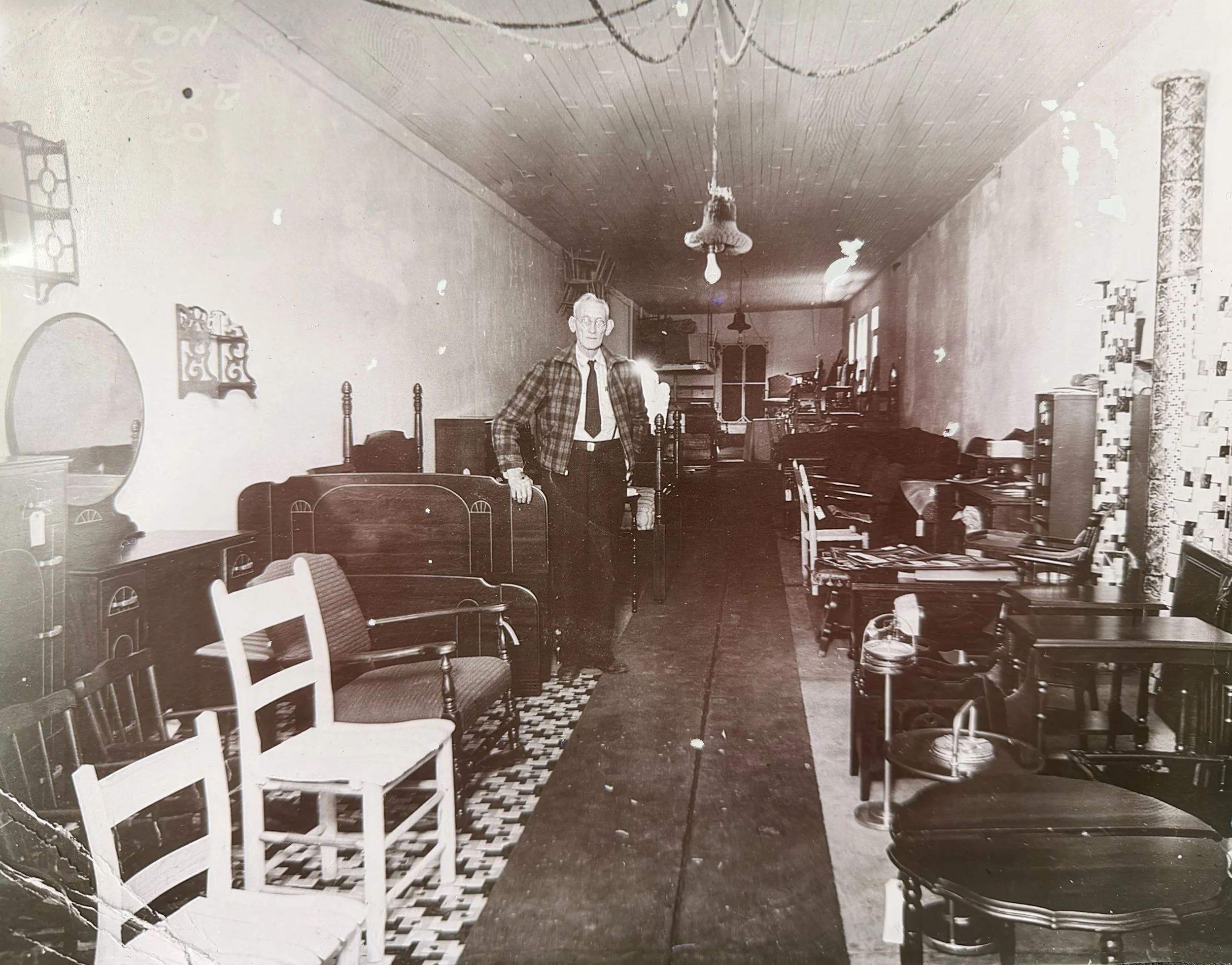 A black and white photo of a man standing in a vintage room filled with furniture and decor. The man is dressed in a plaid shirt, tie, and dark pants, standing next to a bed and surrounded by chairs, tables, and various household items.