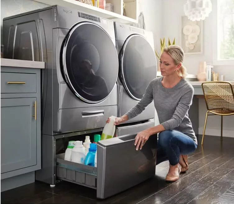A woman is crouching down and opening the bottom drawer of a laundry machine filled with various cleaning supplies in a laundry room.