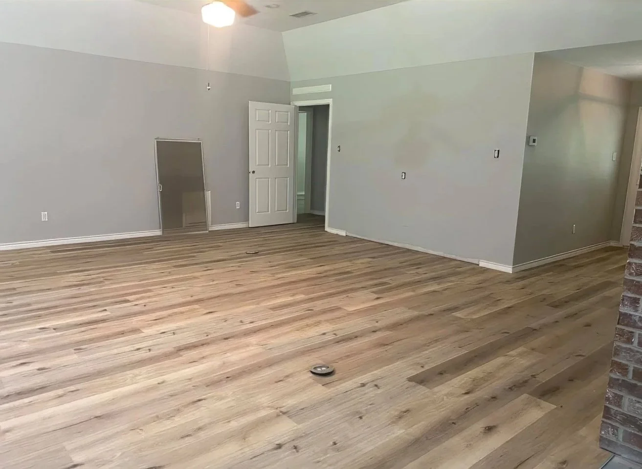 Empty living room with new light-colored wooden flooring, gray walls, ceiling fan with lights, and a partially open door leading to another room.