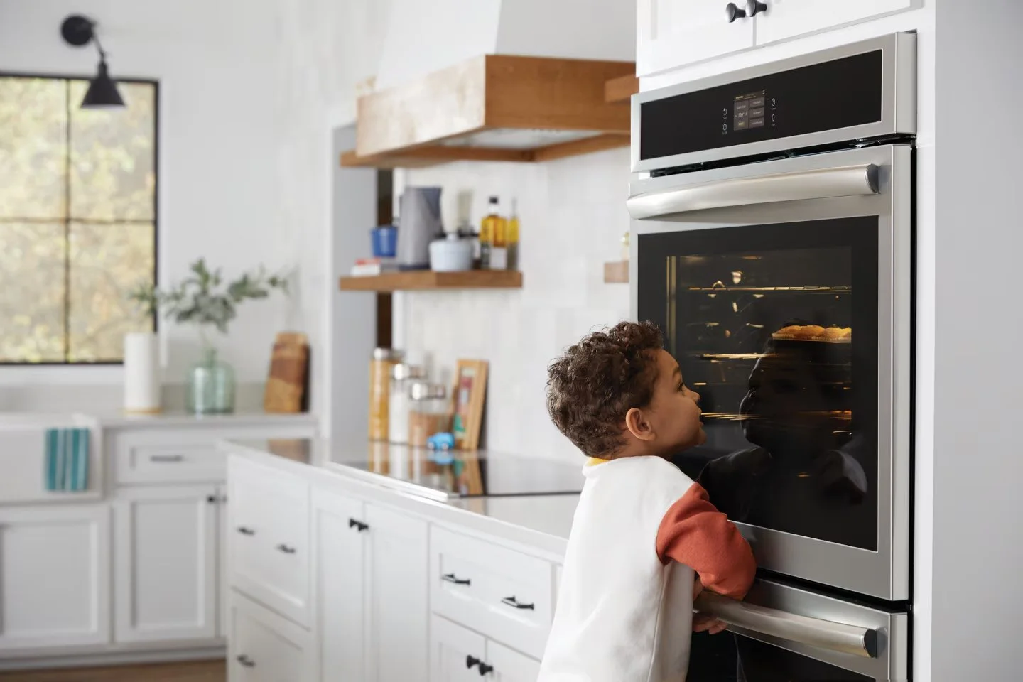 A young child with curly hair looking into an oven in a bright, modern kitchen.