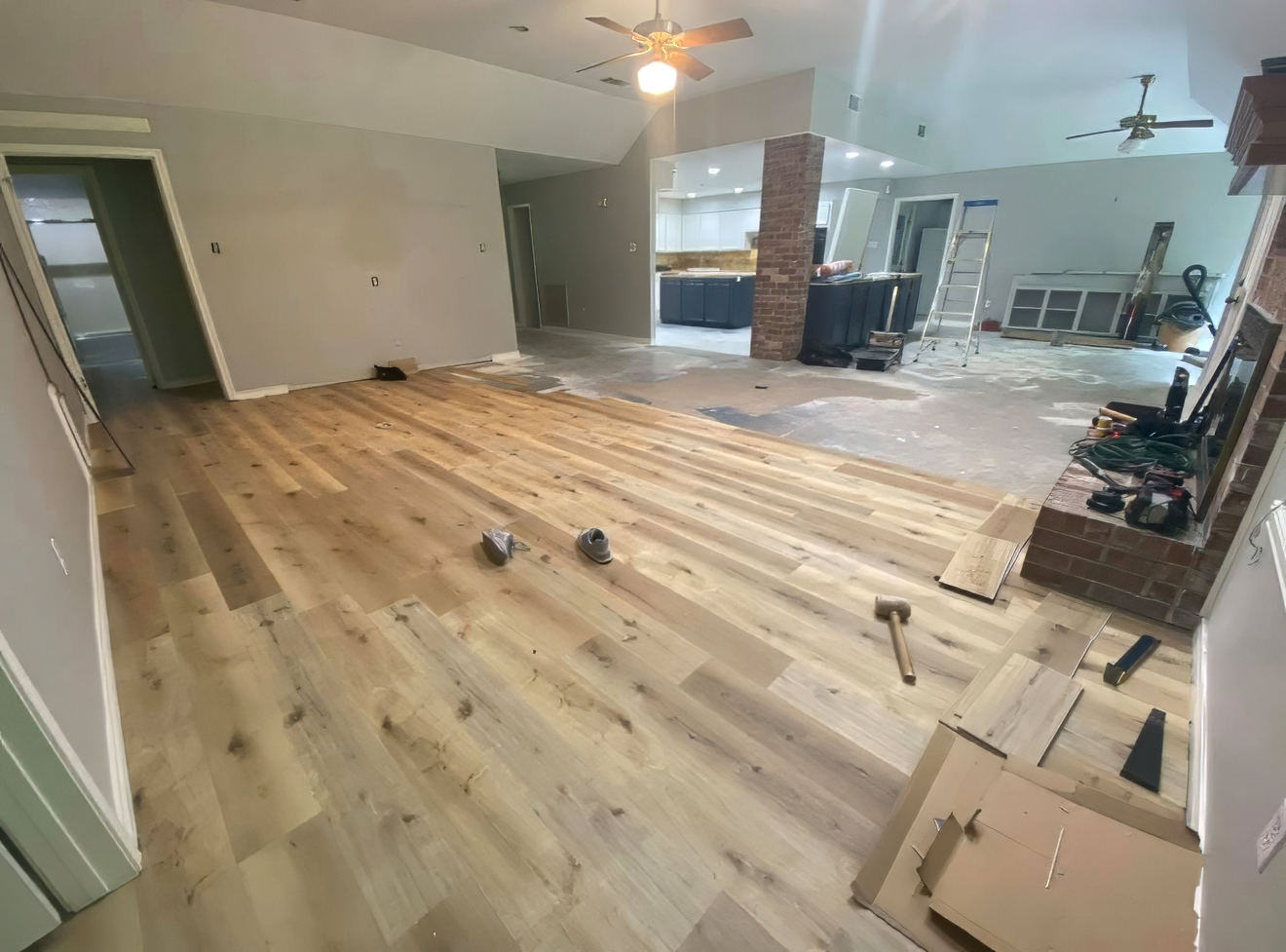Living room with hardwood flooring installation in progress, tools scattered around, and the kitchen visible in the background with brick pillars and a ladder.