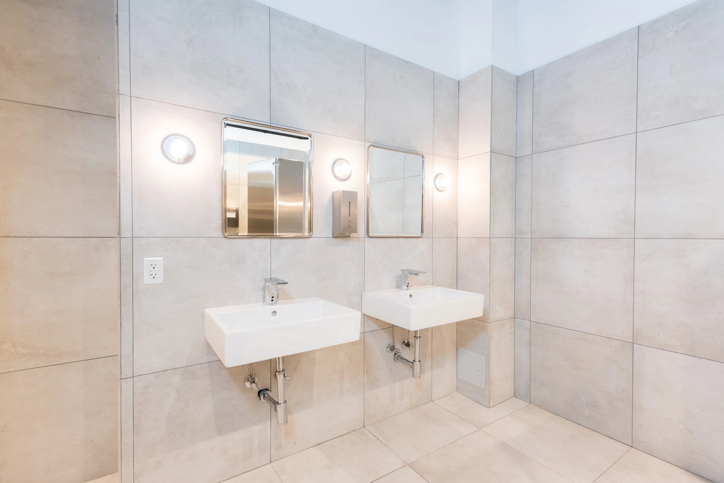 Two wall-mounted sinks with mirrors and light fixtures in a clean, modern bathroom with beige tiled walls.