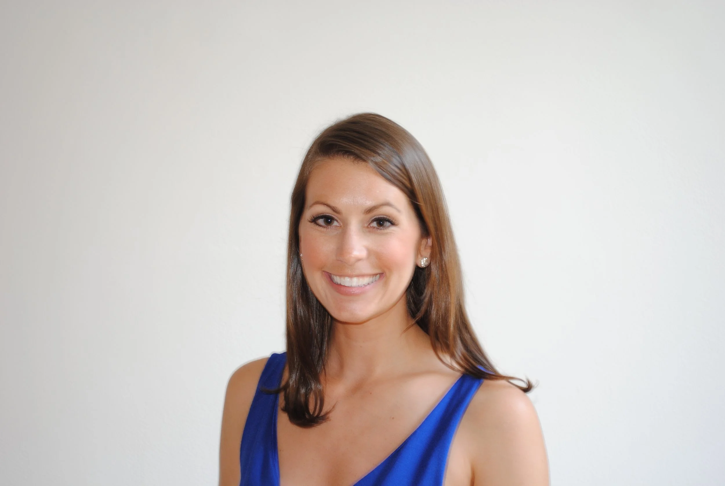 A woman with brown hair, smiling, wearing a blue sleeveless top, standing against a plain white background.