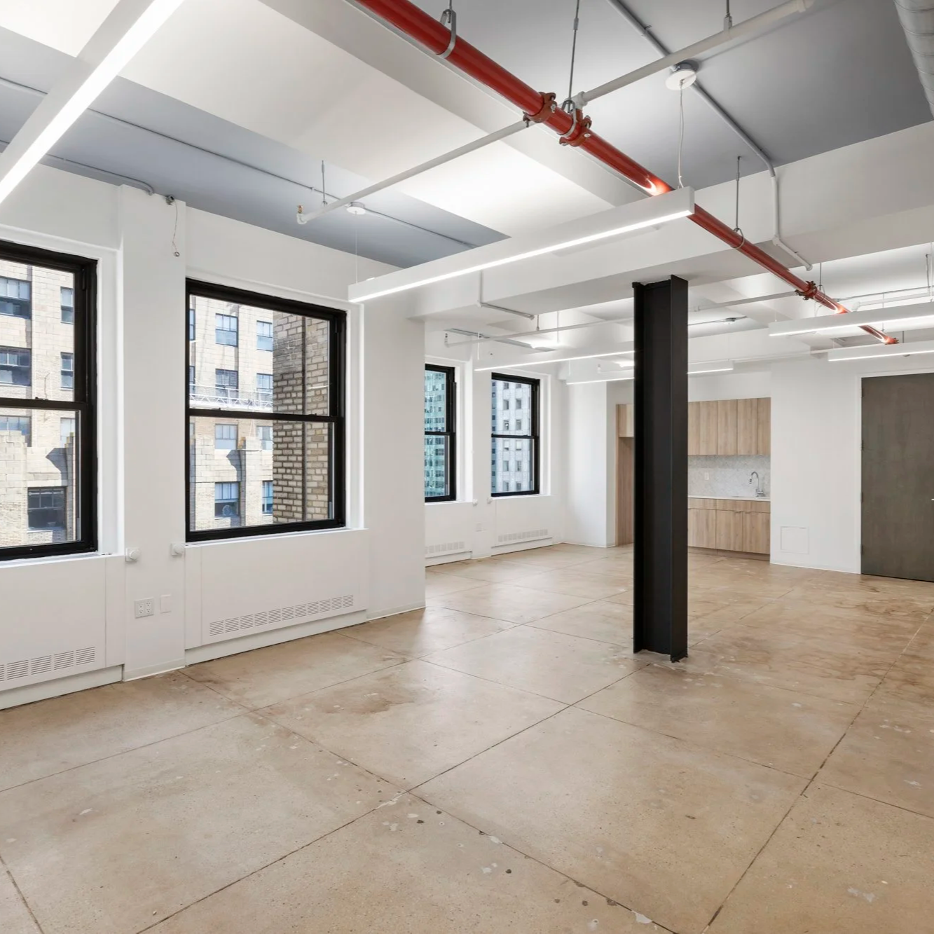 Empty modern  office space with large windows, exposed ceiling pipes, and a kitchenette in the corner.