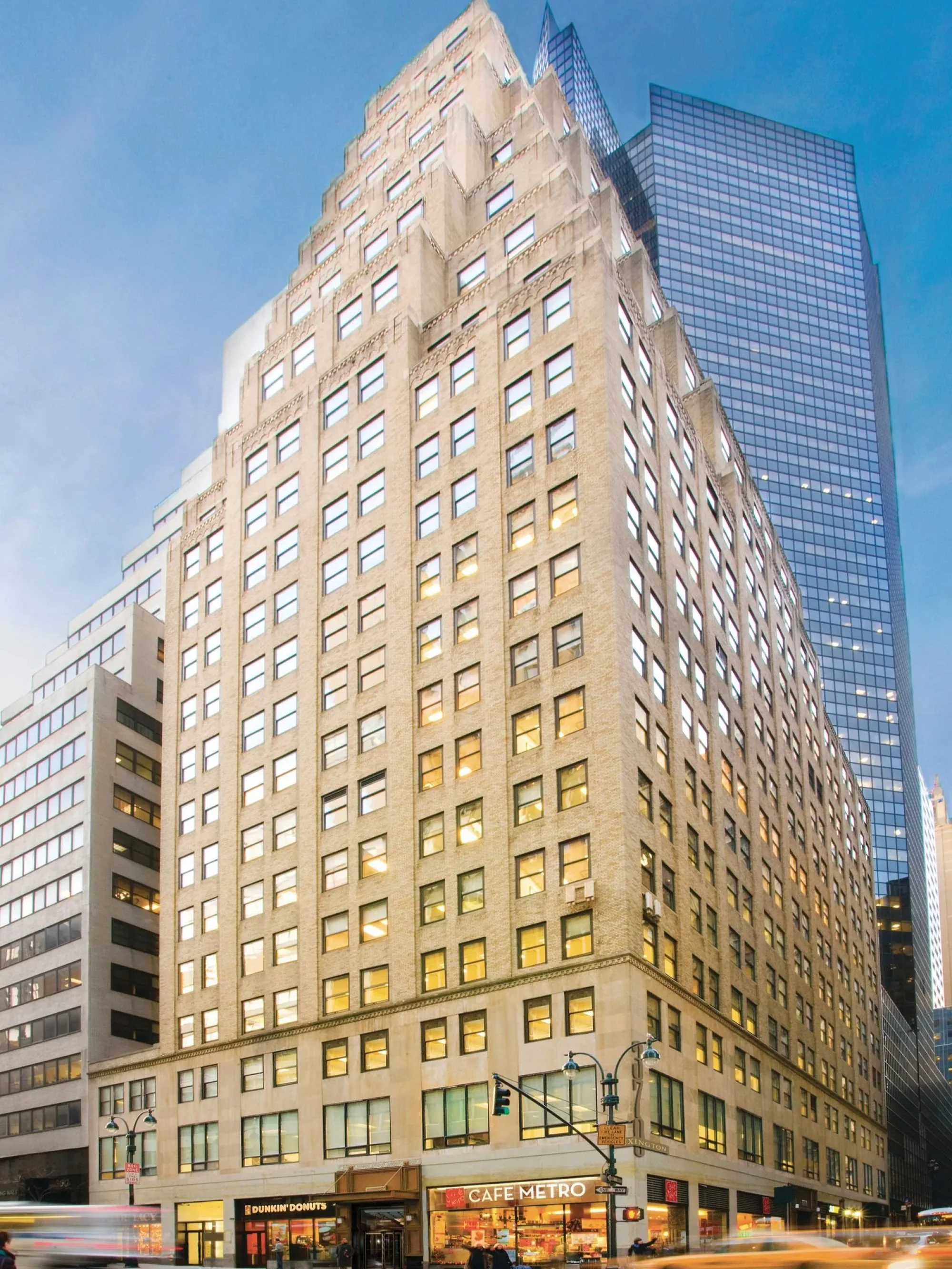 Tall beige building (370 Lex) with numerous windows, street-level shops including Dunkin' Donuts and Le Pain Quotidien, Zuckers, and traffic lights, set against a backdrop of modern glass skyscrapers in a city.