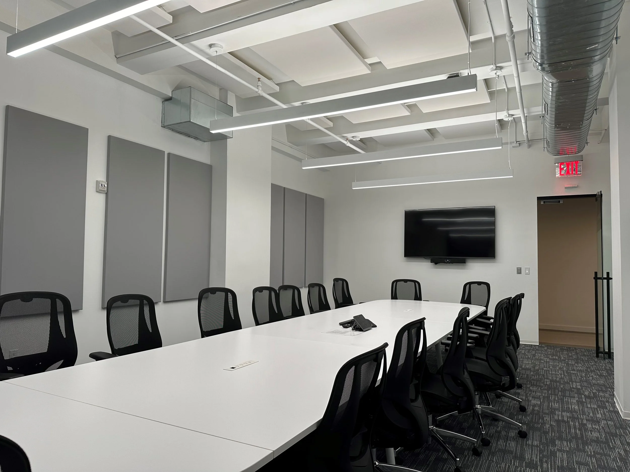 Modern conference room with a long white table, black mesh office chairs, a wall-mounted TV, and acoustic panels on the wall.