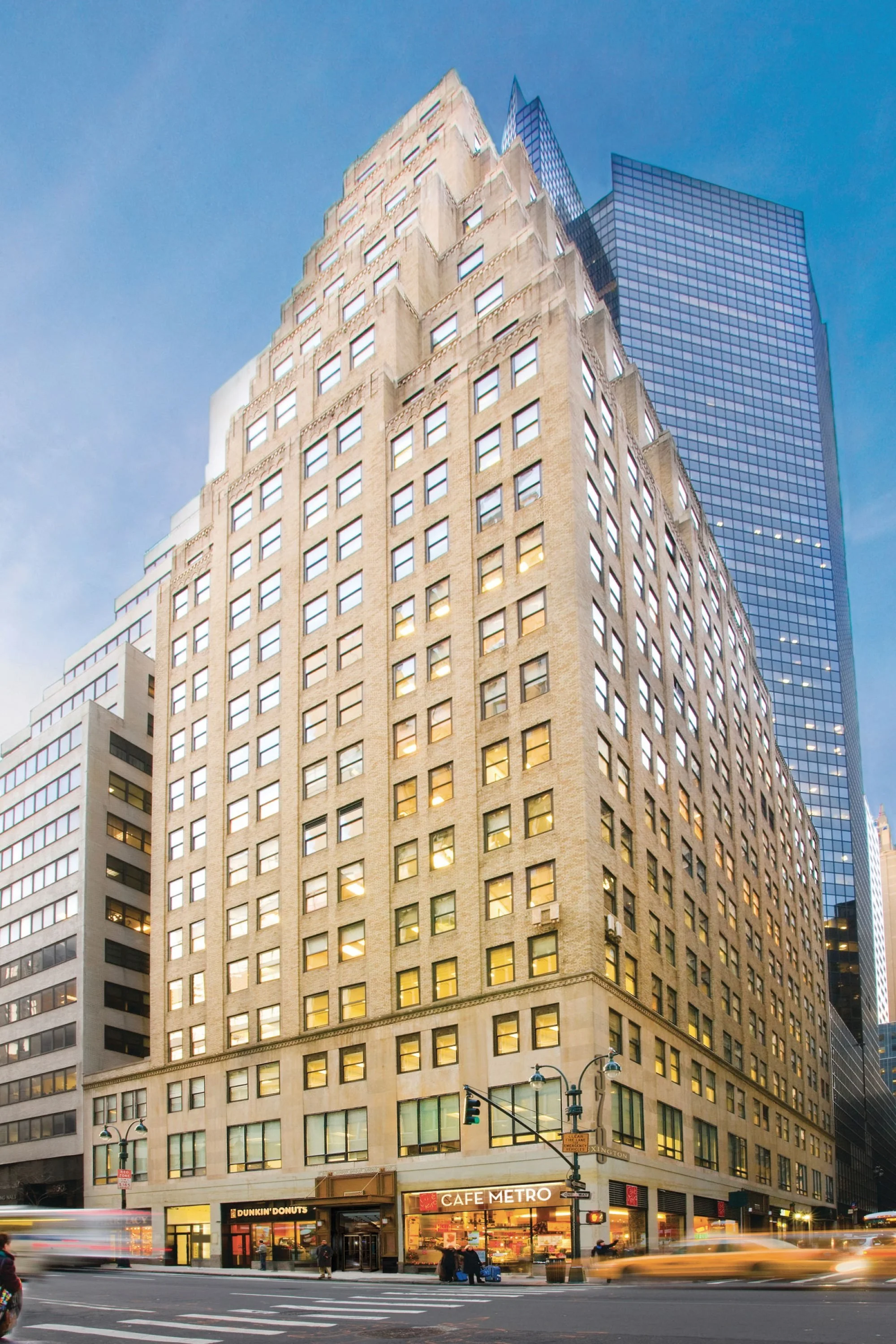 A tall city building with many windows (370 Lexington) with a cafe and donuts shop at the ground level, and a blue skyscraper behind it.
