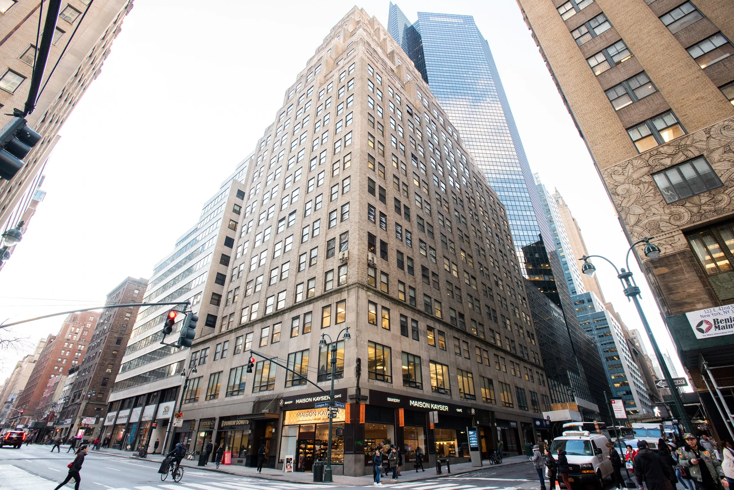 A busy city street with tall buildings, people walking and cycling, and traffic lights in the foreground. A bakery named Maison Kayser is located at the corner of the street.