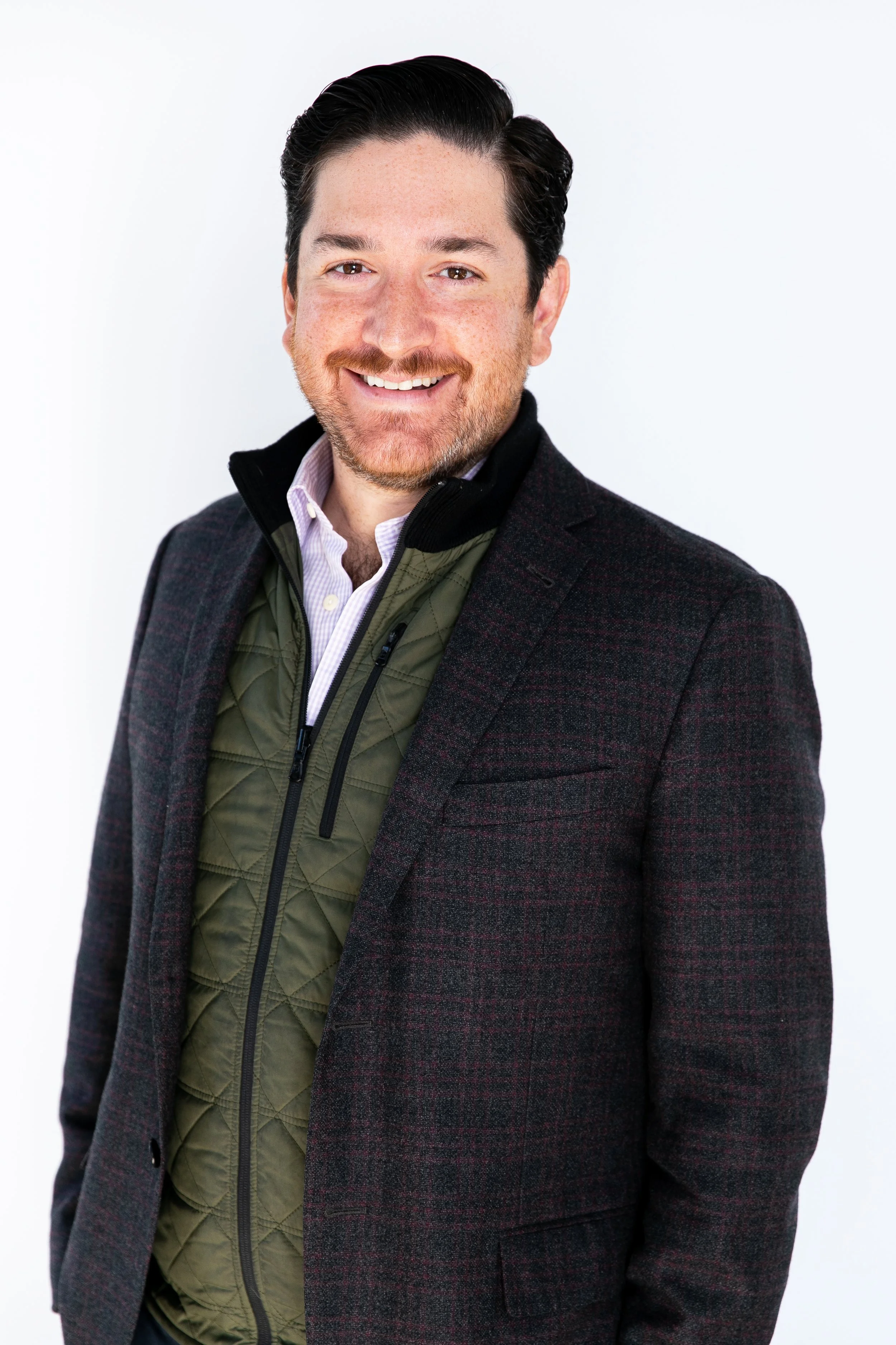 A smiling man with dark hair, beard, and mustache, wearing a dark blazer over a green quilted vest and a light-colored dress shirt, standing against a plain white background.