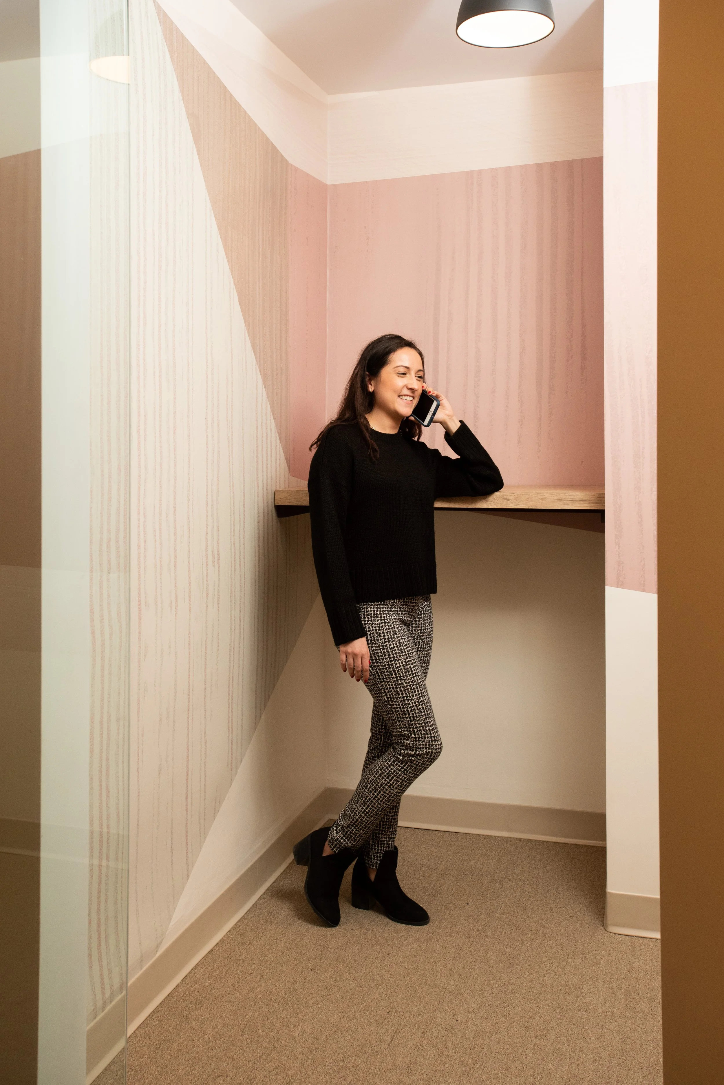 A woman standing in a corner of a room, smiling and talking on a cell phone, wearing a black sweater, patterned pants, and black ankle boots.