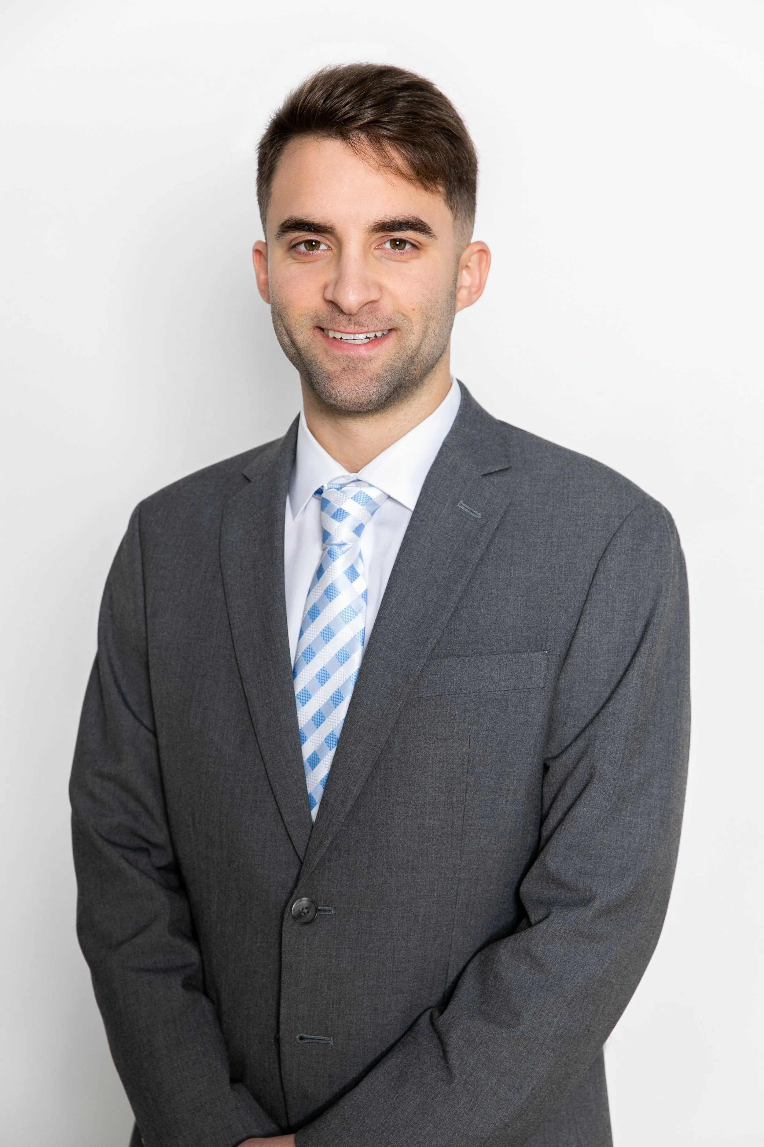 A young man with dark hair and a light beard, wearing a gray suit and a light blue plaid tie, standing against a white background and smiling.