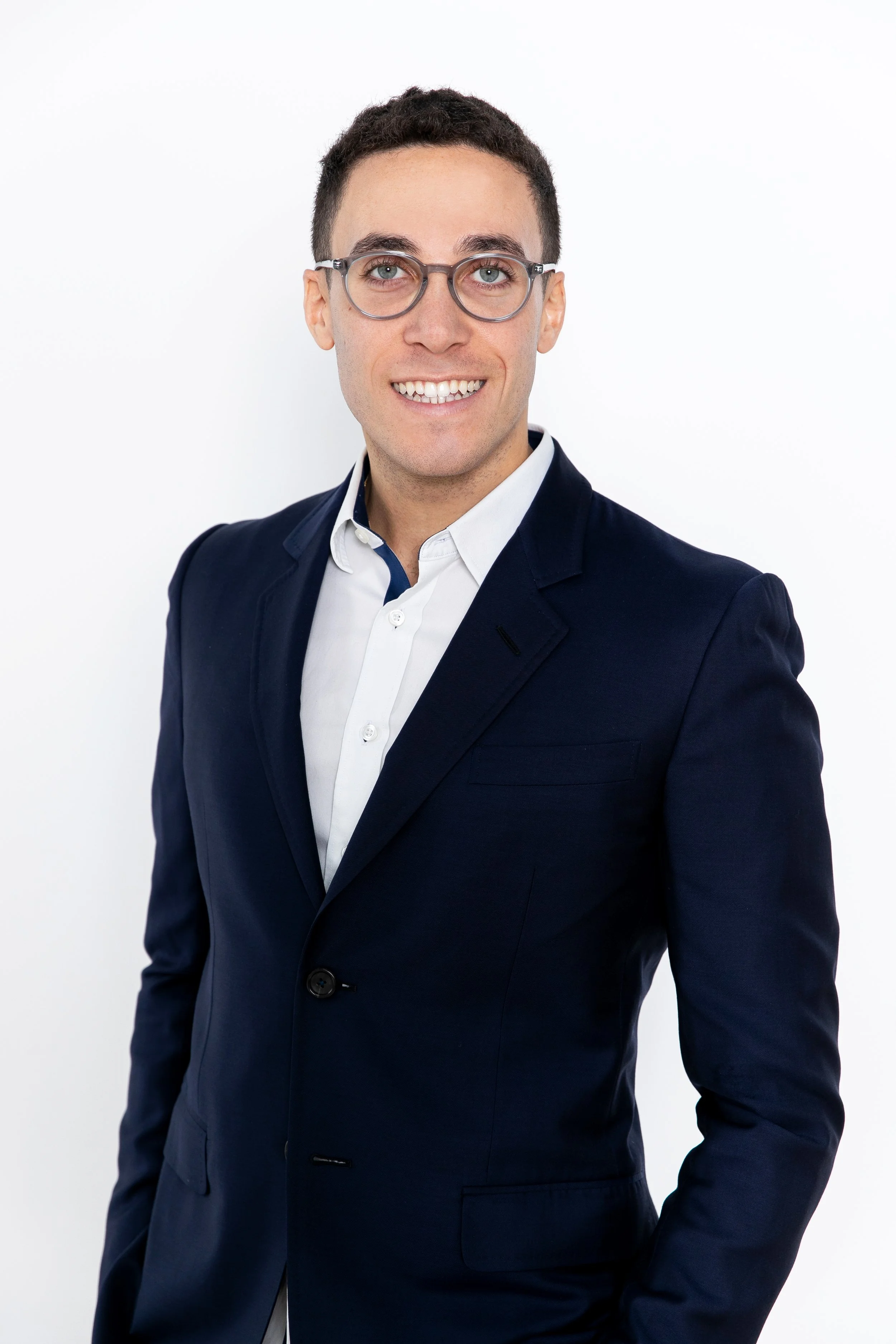 A young man in a dark suit with a white shirt, wearing glasses, smiling, against a plain white background.