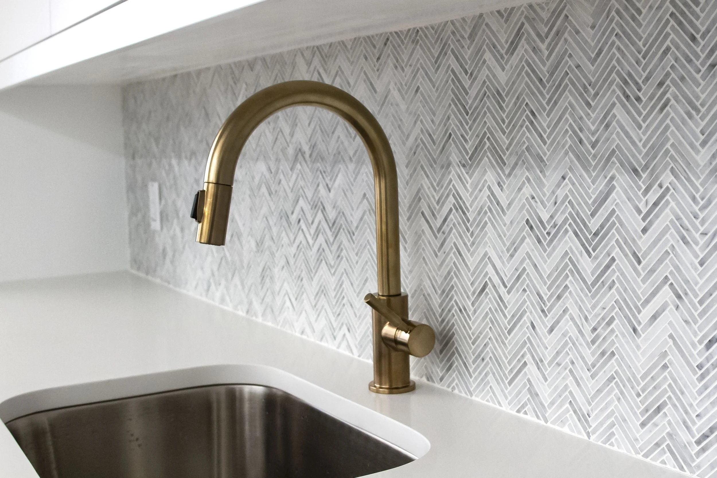 Close-up of a modern kitchen faucet with a gold finish mounted on a white countertop. The background features a textured tile backsplash with a chevron pattern in light gray and white.