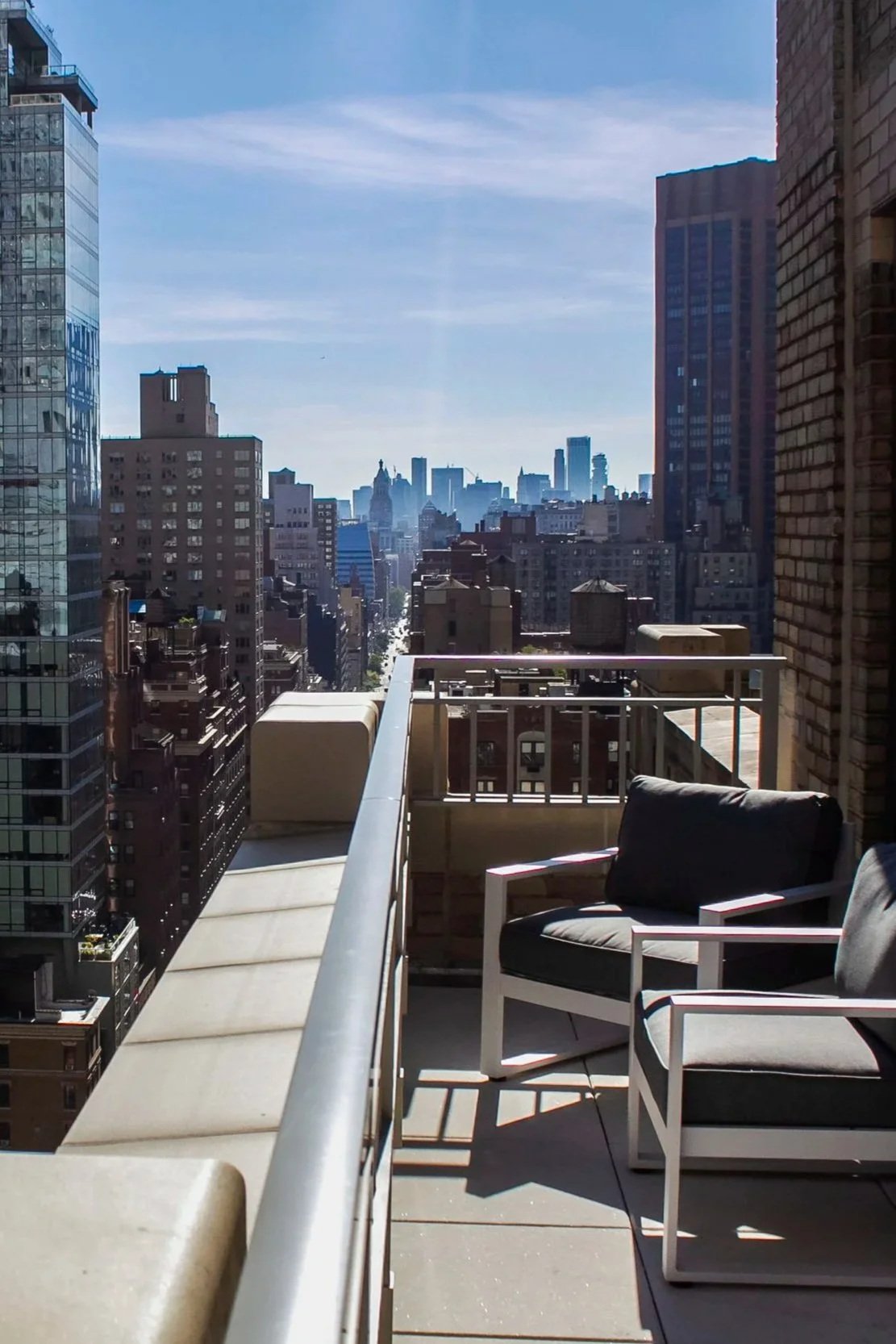 Balcony with two black cushioned chairs overlooking a city skyline with tall buildings and blue sky.