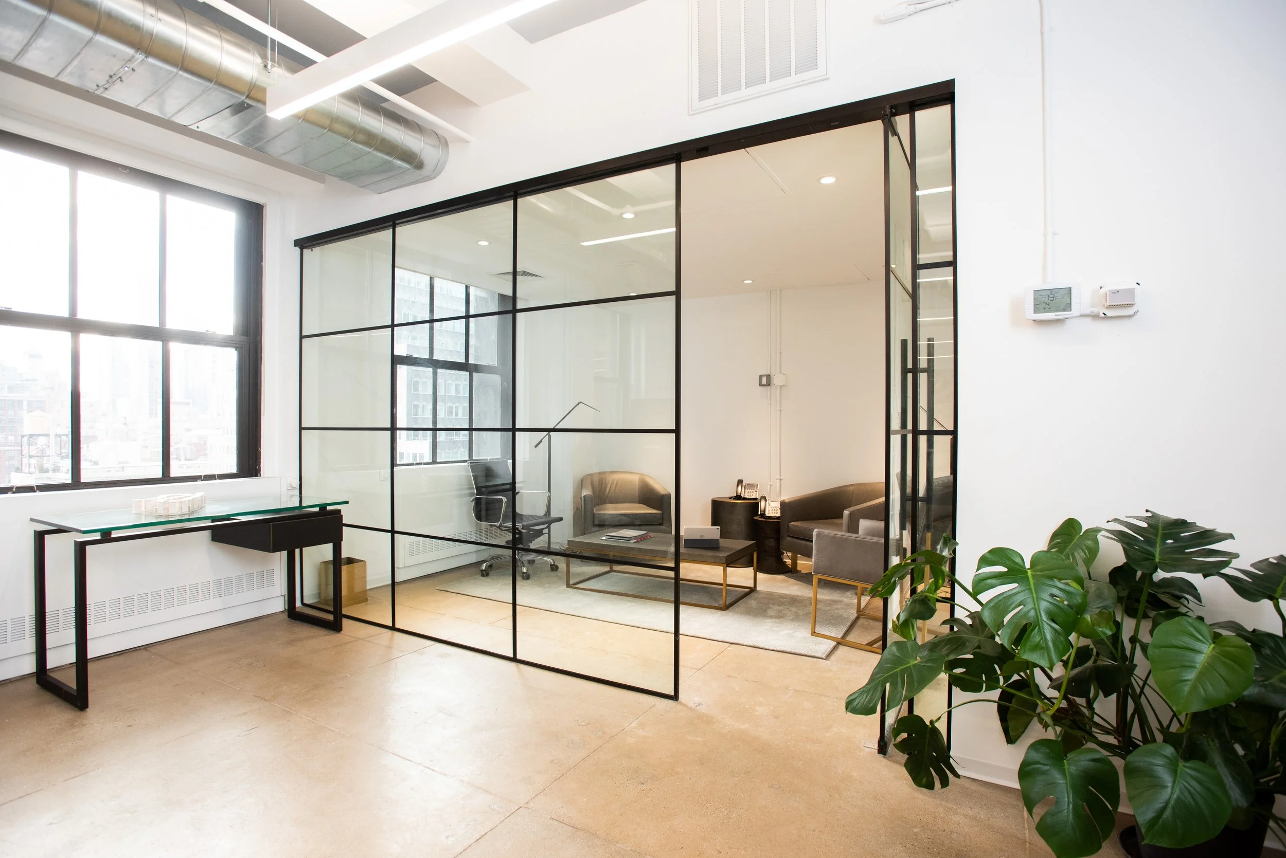 Modern office interior with glass partition, seating area with chairs and a coffee table, large window, and potted plant.
