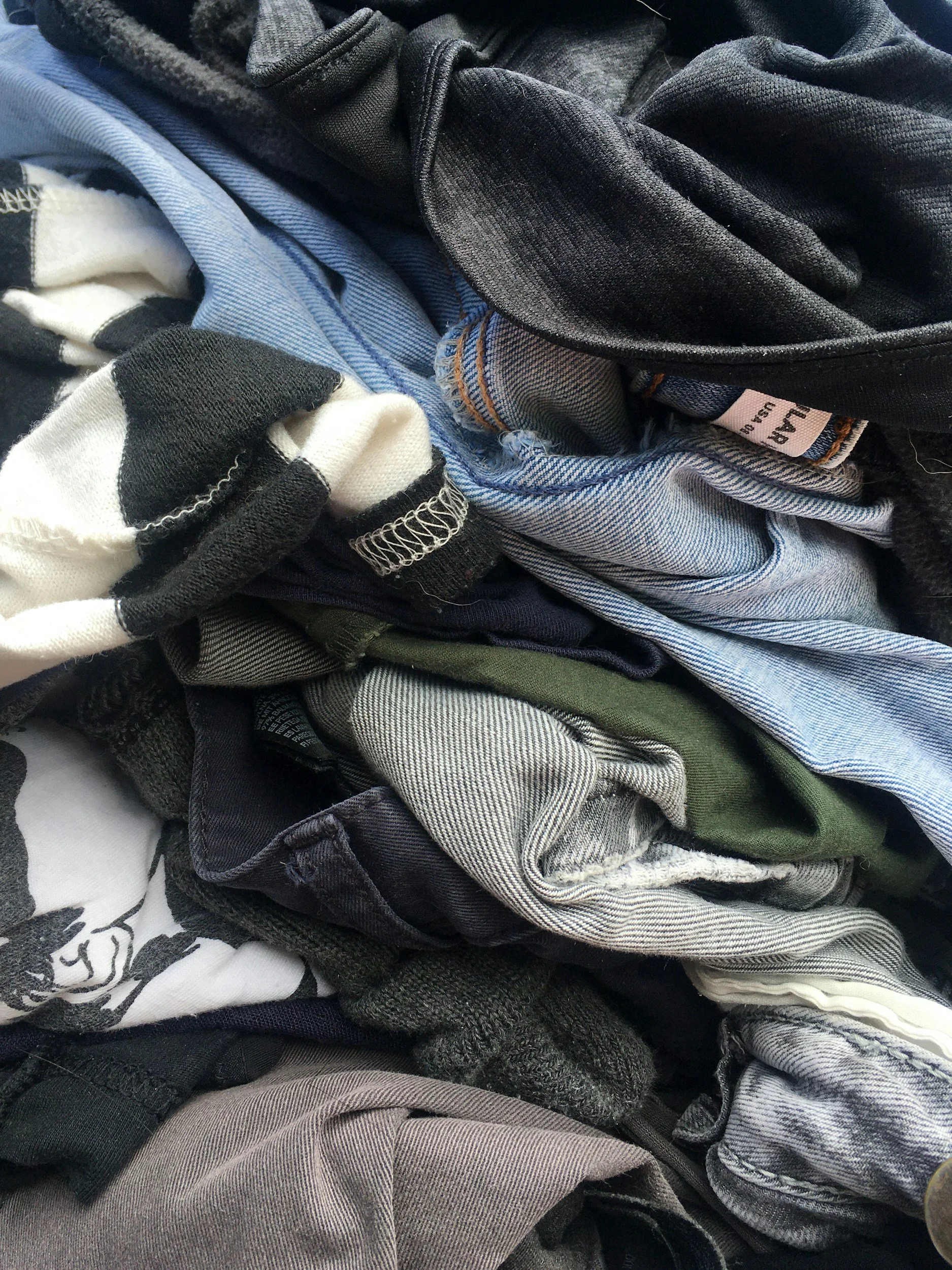 A pile of assorted clothing including jeans, shirts, and fabric in shades of black, gray, blue, white, and green.
