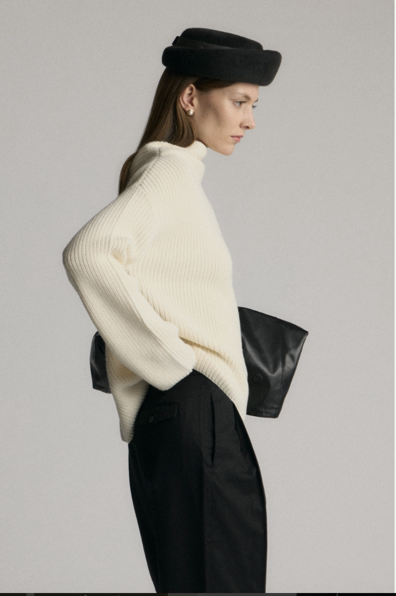 Side view of a woman wearing a black hat, cream-colored turtleneck sweater, and black pants, holding a black clutch bag.
