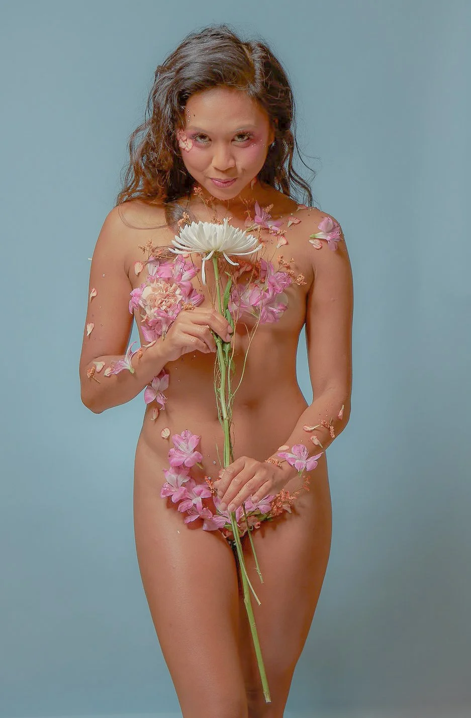 A nude woman with pink and white flowers and petals on her body, holding a white flower, standing against a light blue background.