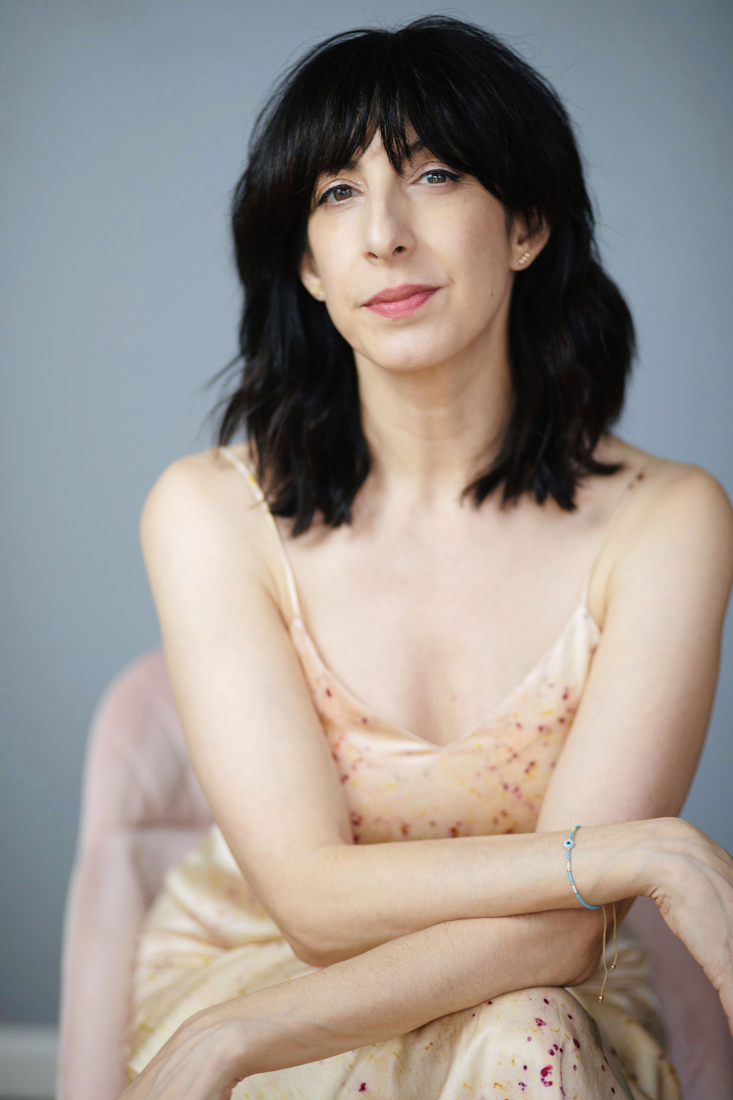 A woman with shoulder-length black hair and light skin, wearing a pale yellow dress with pink and red speckles, sitting with her arms crossed.