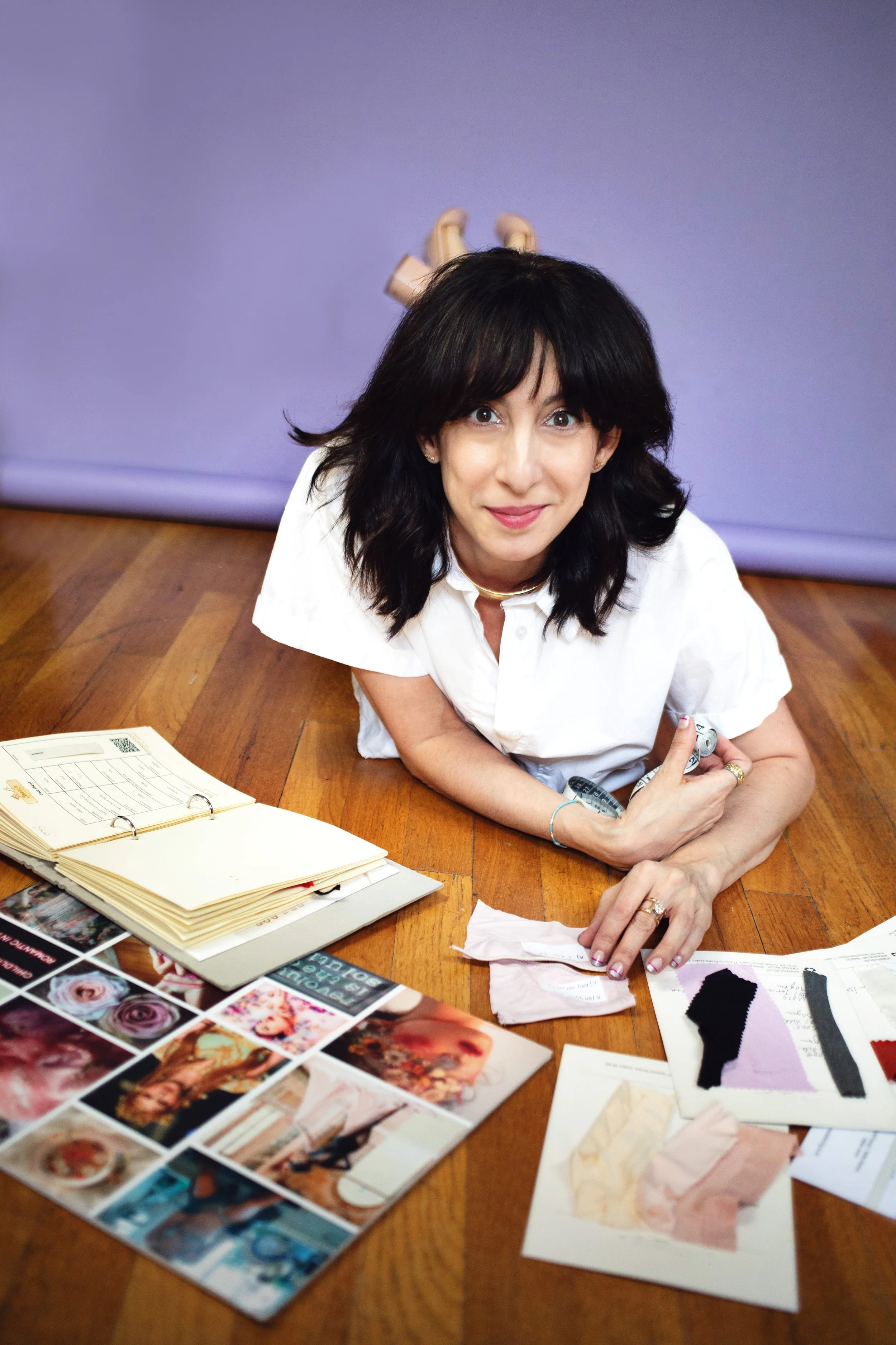 Woman lying on a wooden floor with various papers, fabric samples, and a photo collage spread out in front of her
