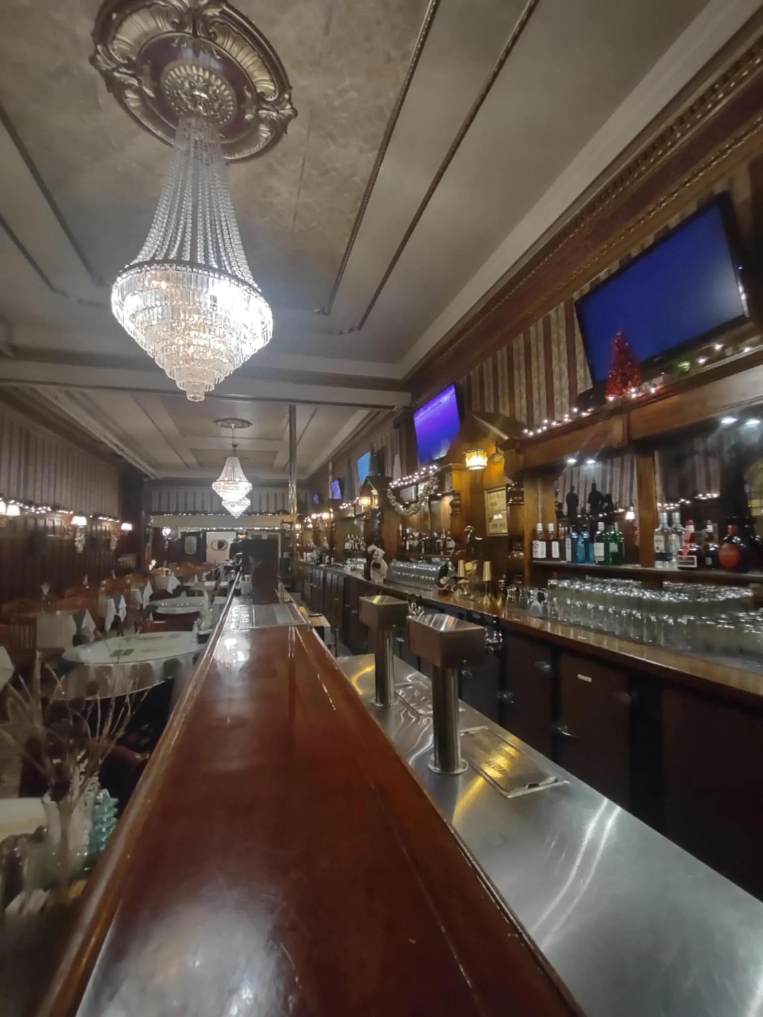 Interior of an elegant restaurant with chandeliers, wooden bar with glasses and bottles, and multiple TV screens on the wall, decorated for Christmas.