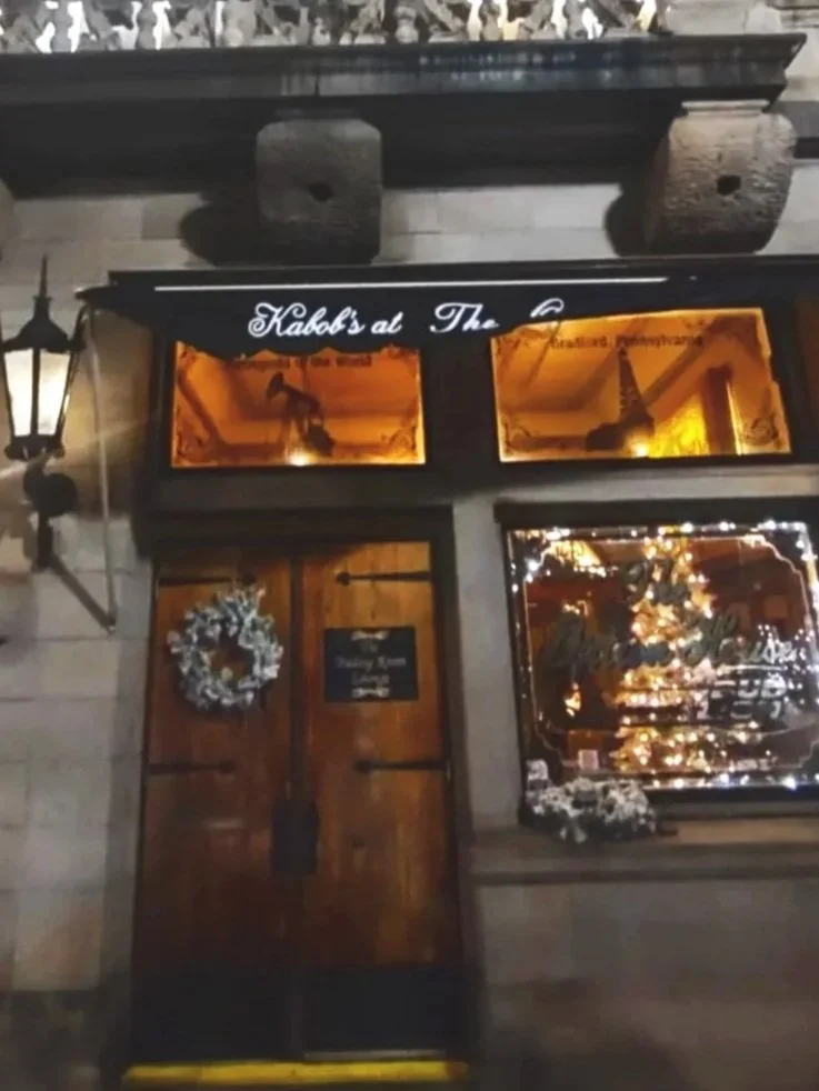 A storefront decorated for the holidays with Christmas lights, a wreath, and a Christmas tree visible through the window. The sign above the door reads "Habb's at The" followed by unreadable text.