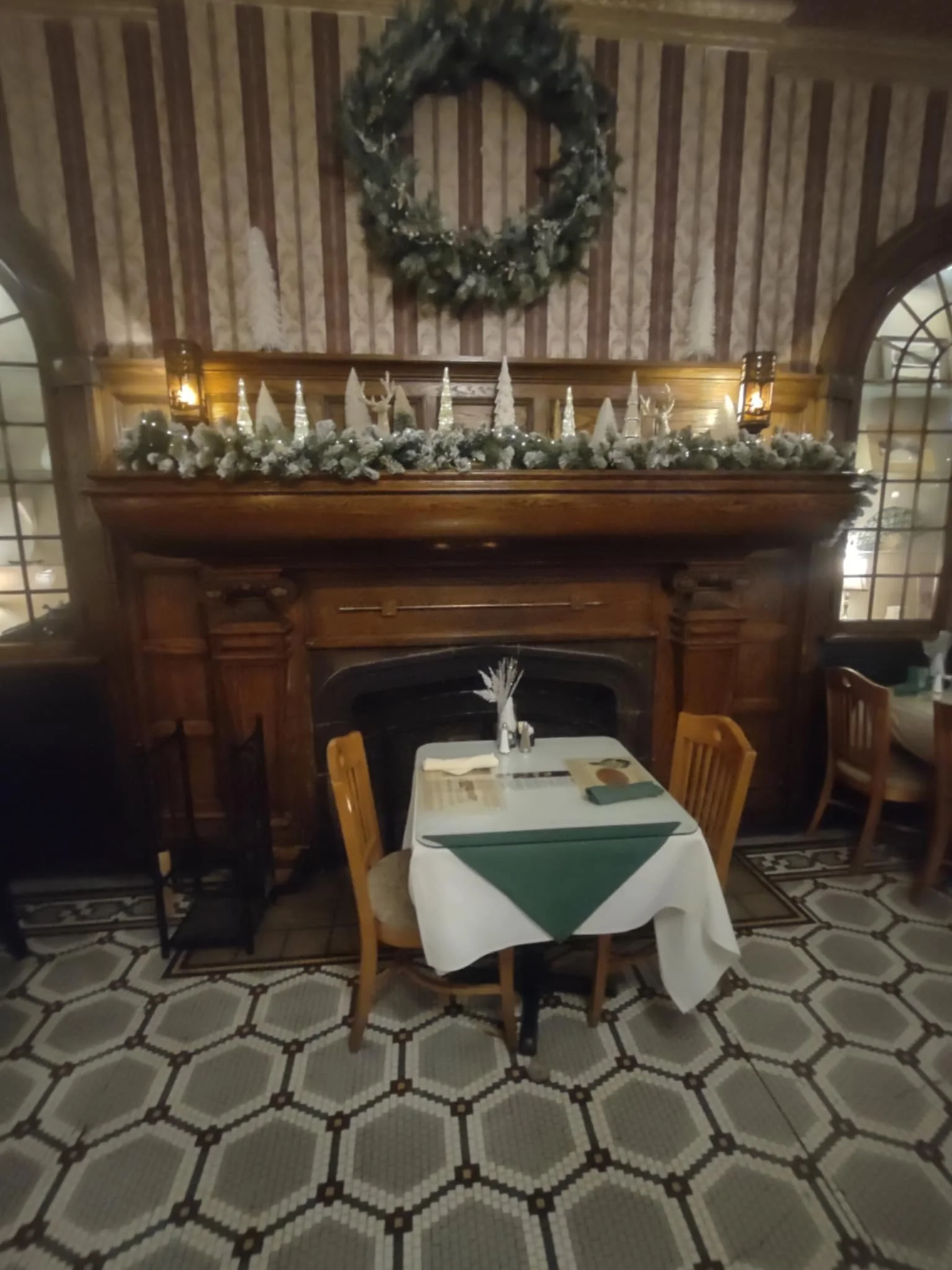 A cozy restaurant setting decorated for Christmas with a fireplace, a wreath hanging above, and a table set with a white tablecloth, green napkins, and festive decorations.