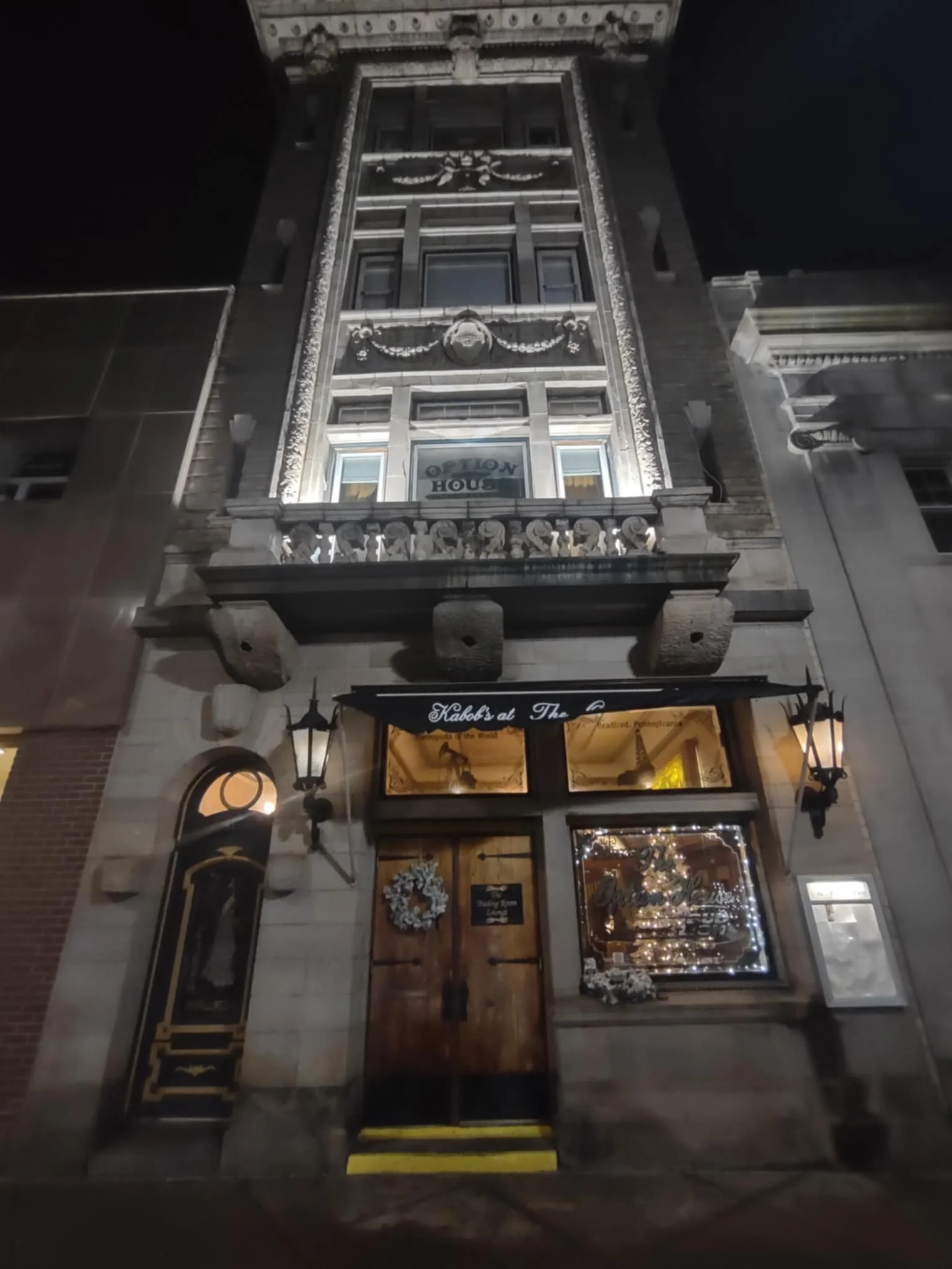 Night view of the historic building with decorative architectural details, illuminated by exterior lights, featuring a wooden door with a holiday wreath and large window with Christmas decorations.
