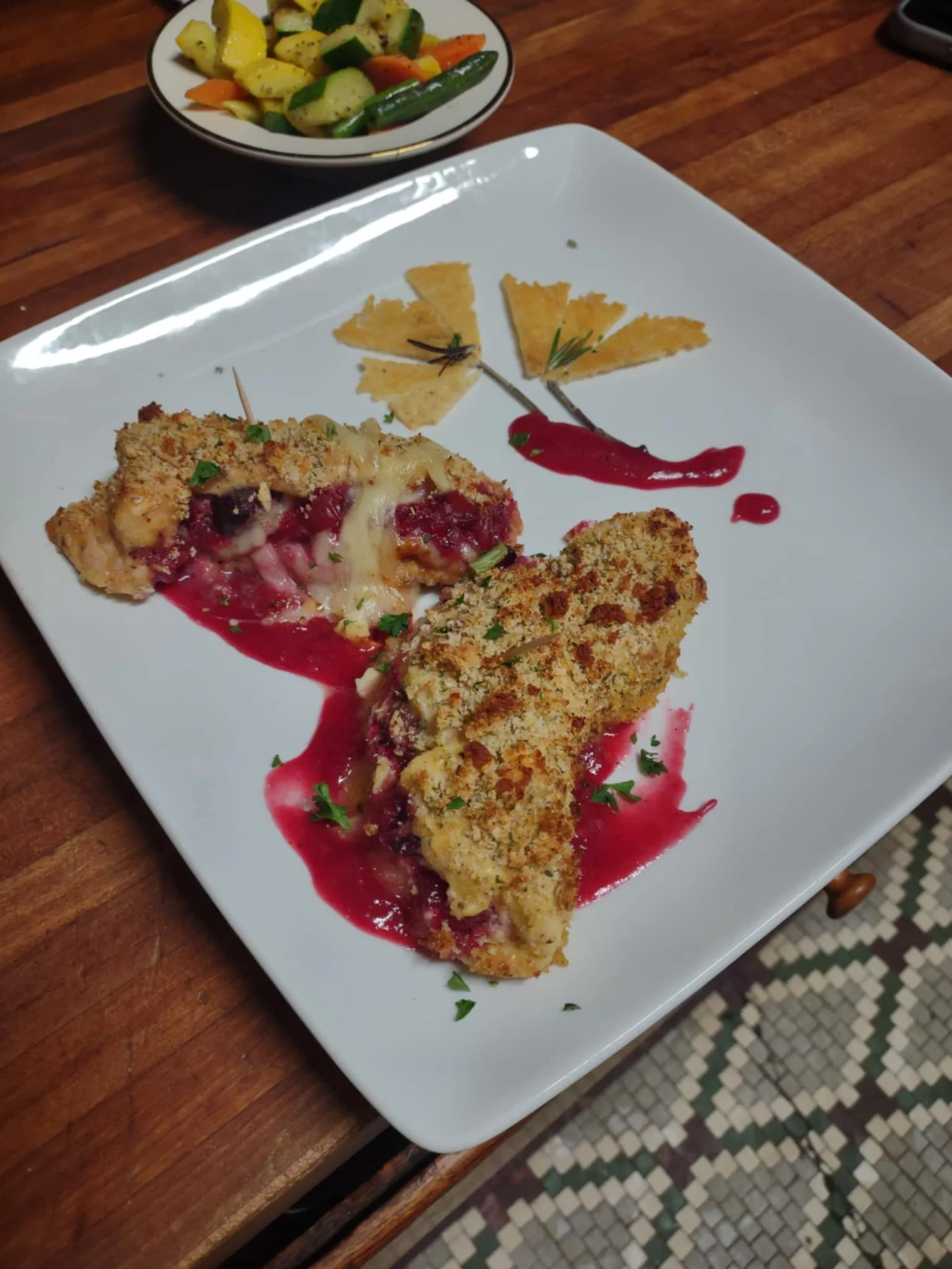 Breaded and baked stuffed chicken breasts with cherry filling, garnished with herbs, served on a white square plate with red sauce, alongside decorative cheese slices with herbs, and a small bowl of sautéed vegetables in the background.