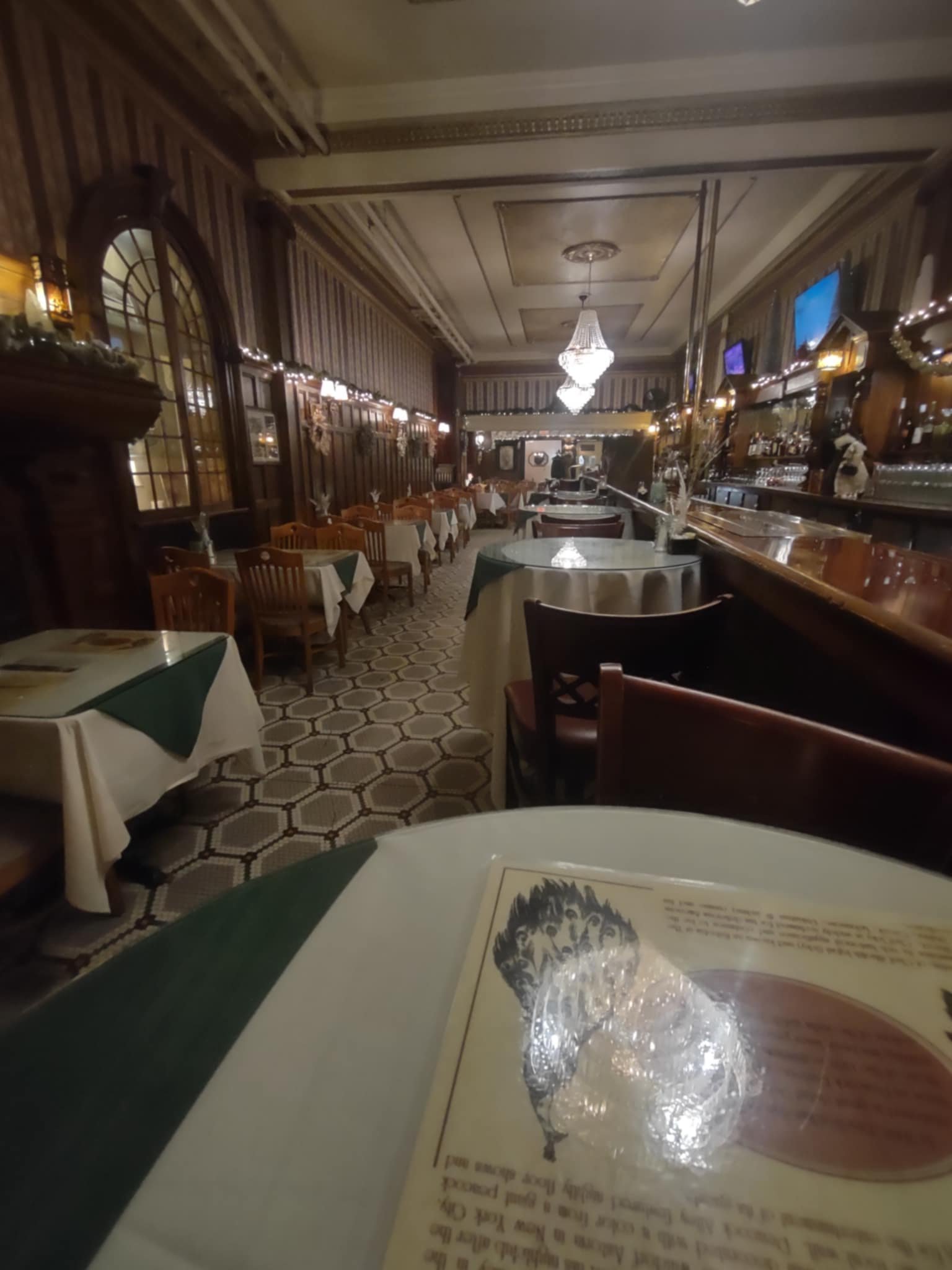 Interior of a vintage-style restaurant with tables covered in white and green tablecloths, a patterned tile floor, wooden chairs, and a bar, decorated with string lights and a chandelier.