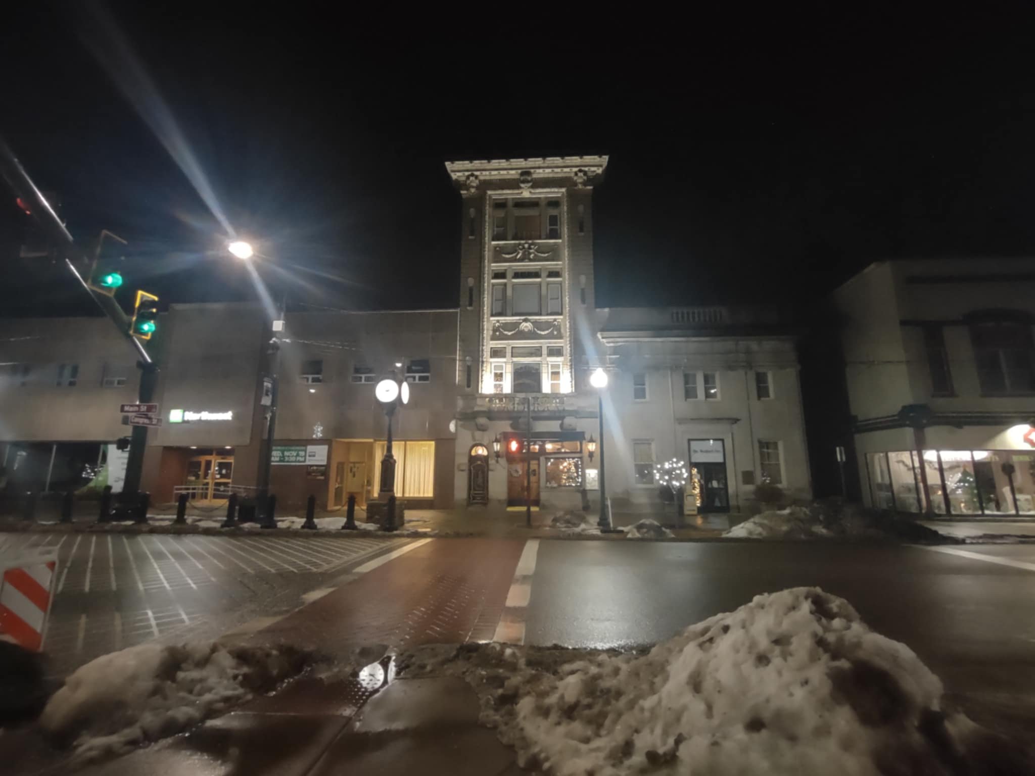 Nighttime street scene with historic building illuminated, traffic lights, snow piles on the sidewalk, and a crosswalk in the foreground.