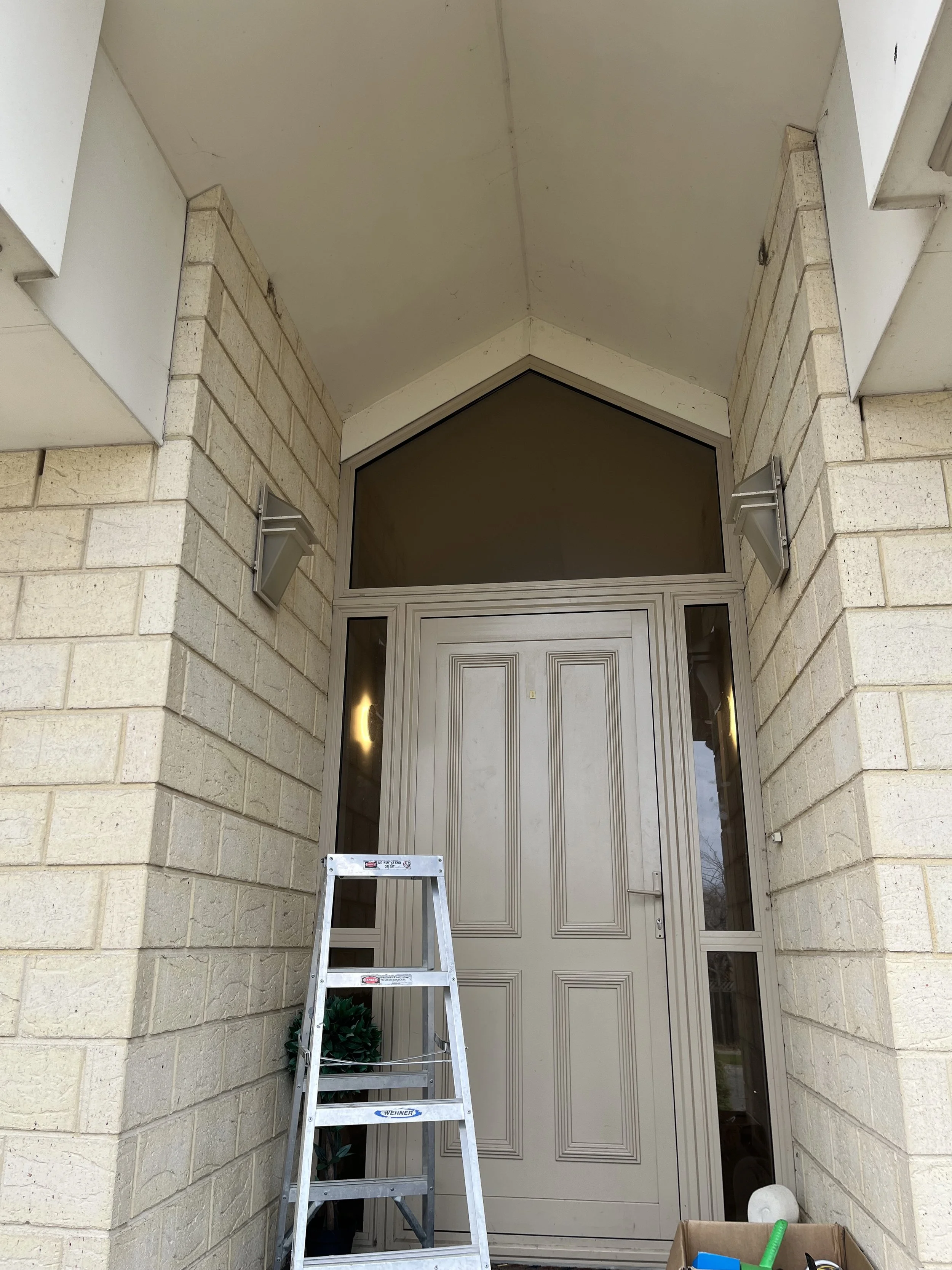 Front entrance with beige door, brick walls, and a step ladder positioned in front of the door with a small potted plant behind it.