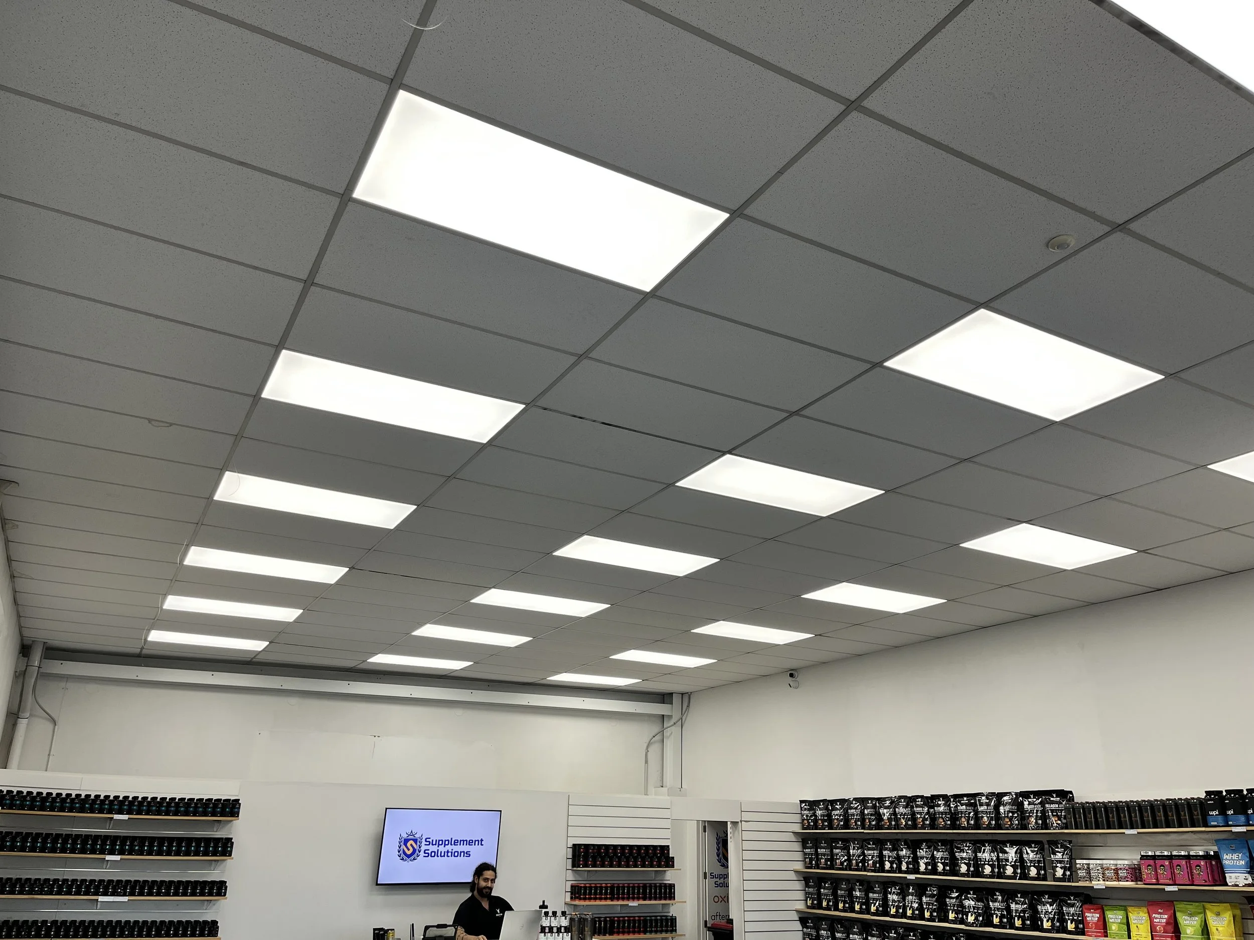 Inside a store selling dietary supplements, with a ceiling of bright rectangular fluorescent lights, shelves filled with protein powder and supplement containers, and a person working at the checkout counter.