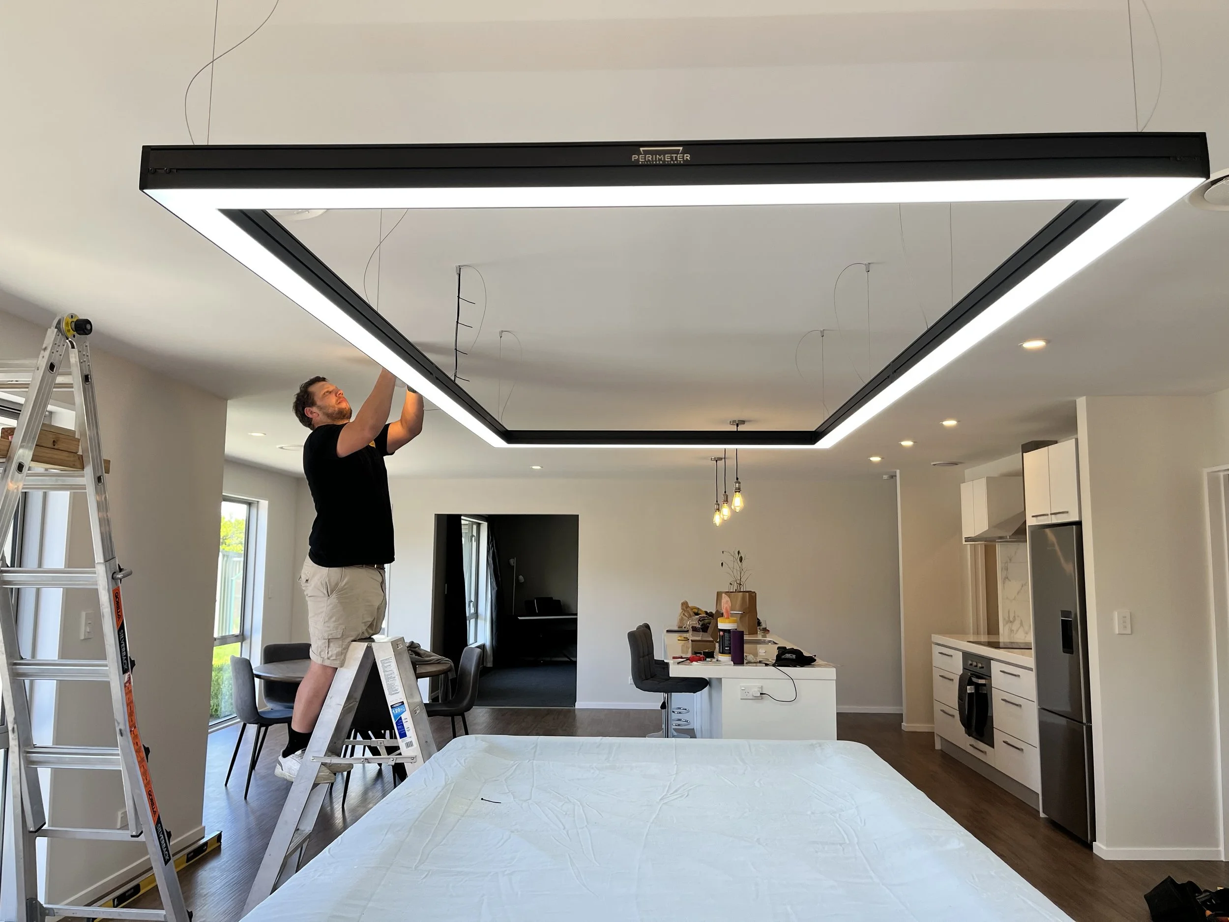 A man standing on a ladder installing or adjusting a large rectangular LED ceiling light fixture in a modern open-plan kitchen and living room.